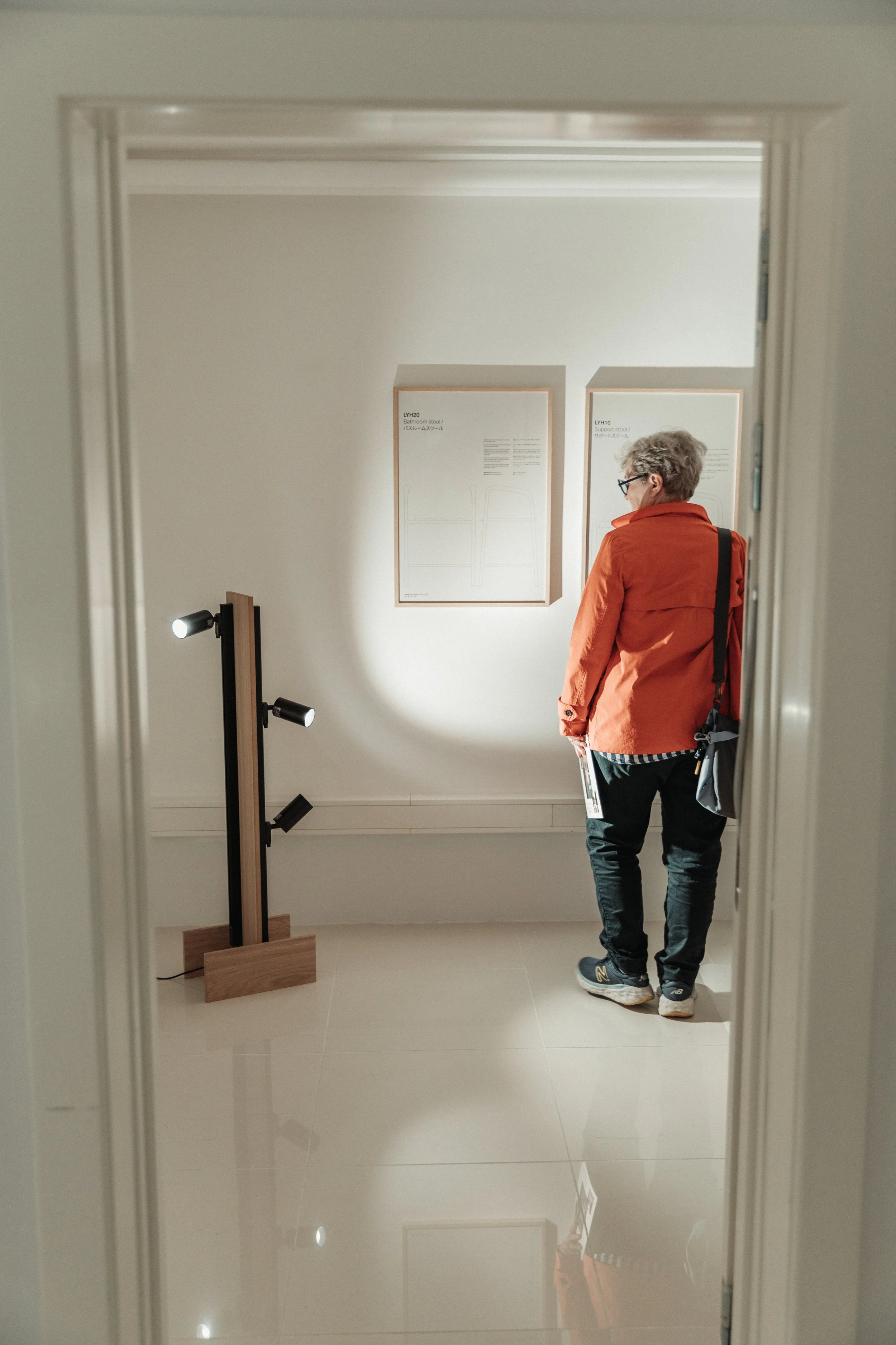 View through a doorway showing a person with gray hair wearing an orange jacket, black pants, and sneakers, standing in front of two framed diagrams labeled 'Bathroom stool' and 'Support stool' on a white wall. To the left, a floor lamp with adjustable lights is visible.