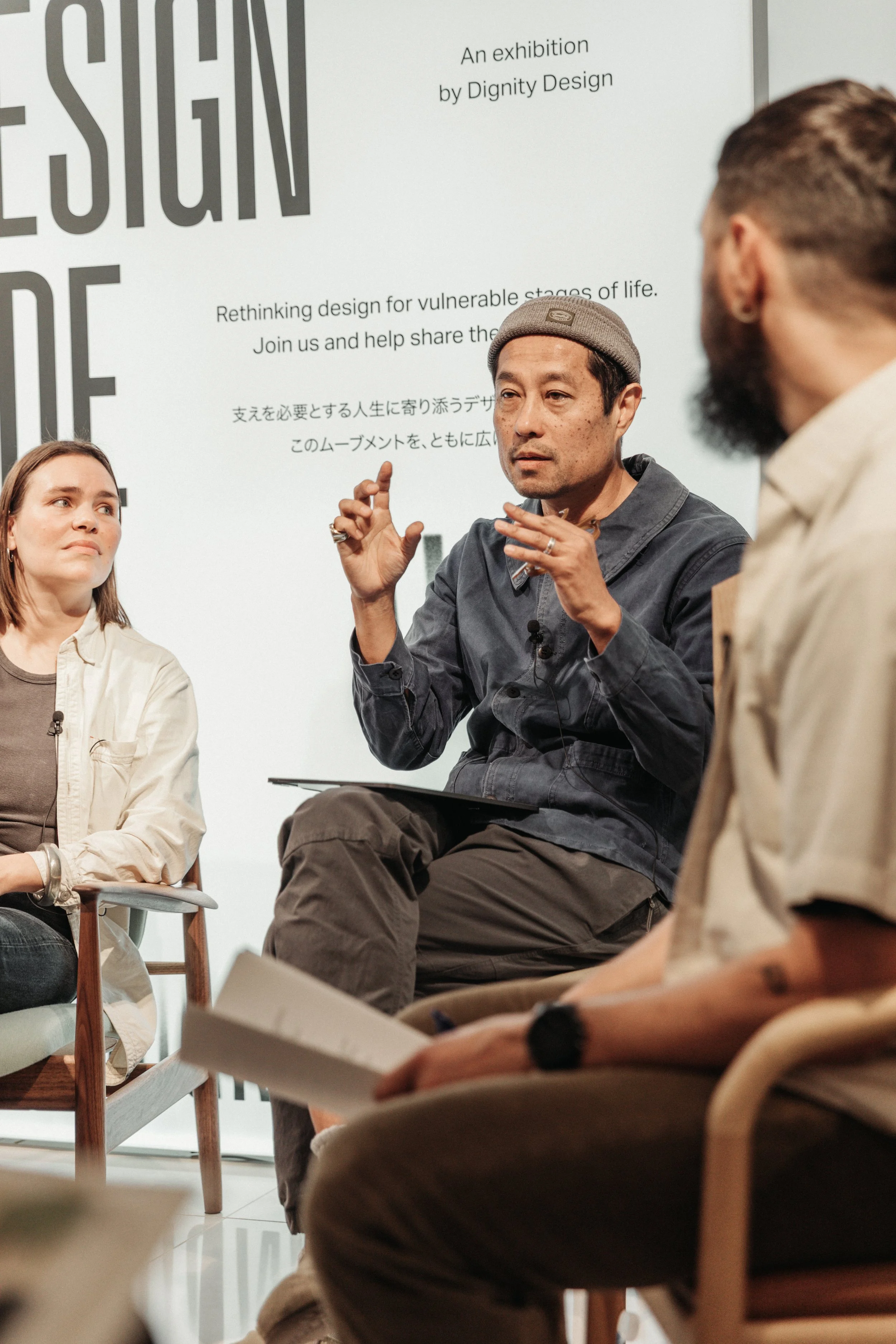 A panel discussion or talk with three people, one man speaking and two women listening, in front of a white wall with text about design and an exhibition by Dignity Design.