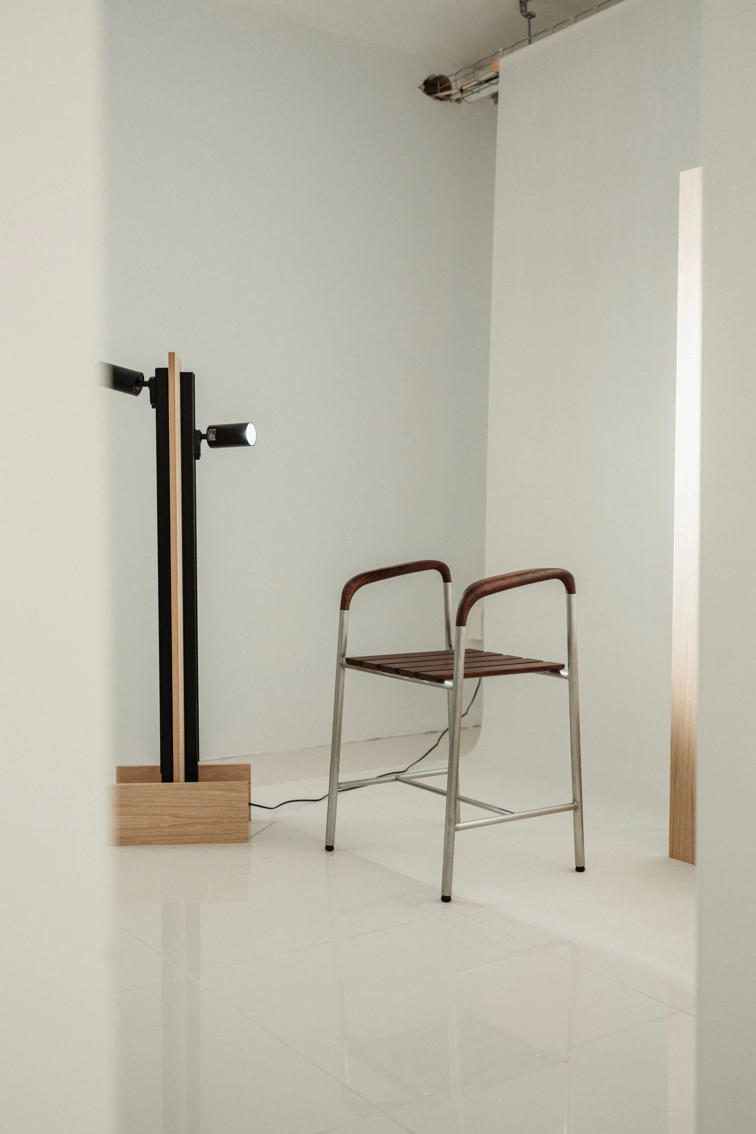 A modern indoor space with a white wall, a wooden floor lamp, a wooden chair with metal legs, and a high ceiling. The floor is white and shiny, and there is a small opening on the wall to the right.