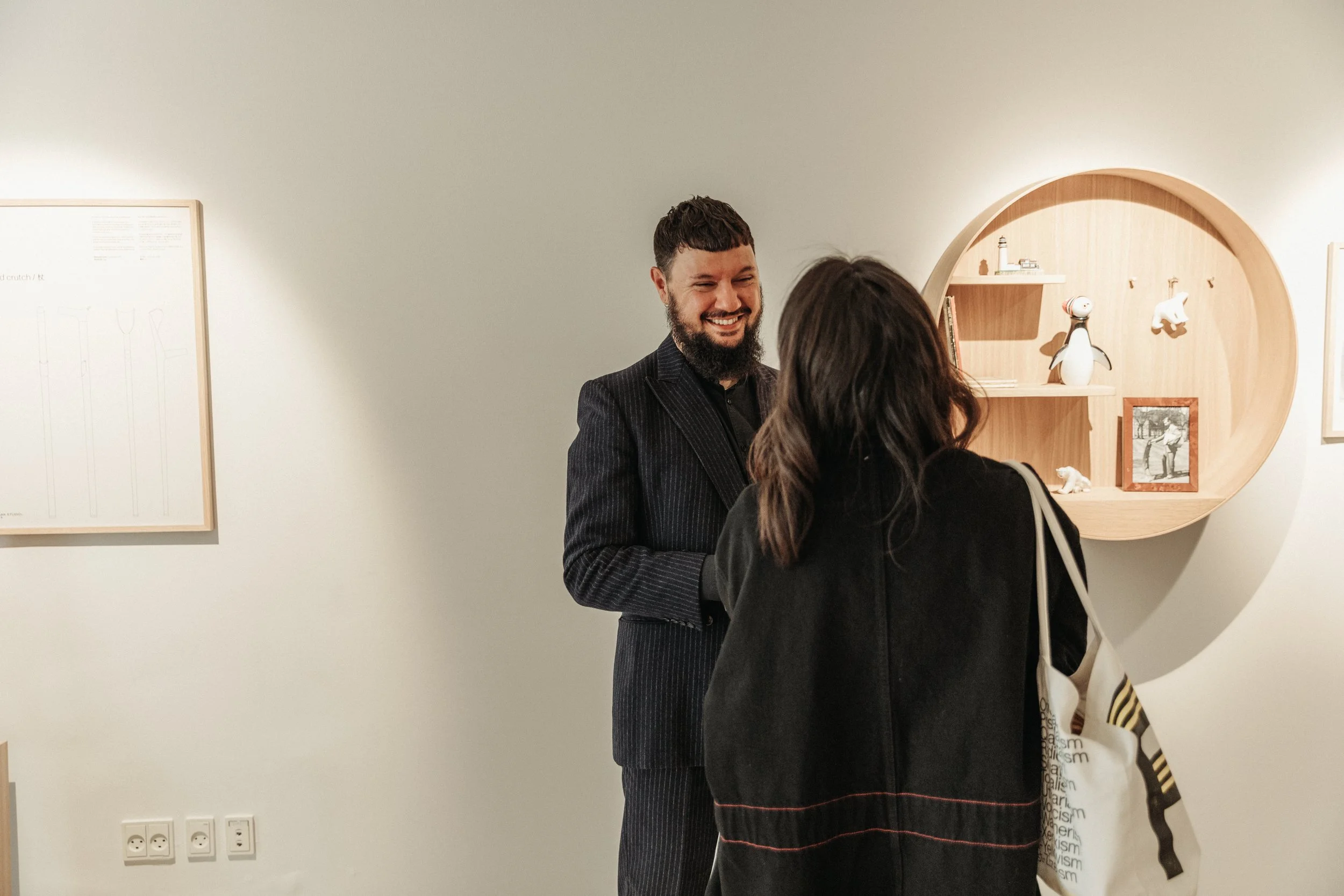Two people, a man with a beard and a woman with long dark hair, are smiling and engaging in a conversation in an art gallery. The wall behind them features a wooden shelf with decorative items and a framed photograph, while to the left there is a white artwork surrounded by a wooden frame and a wall with electrical outlets.