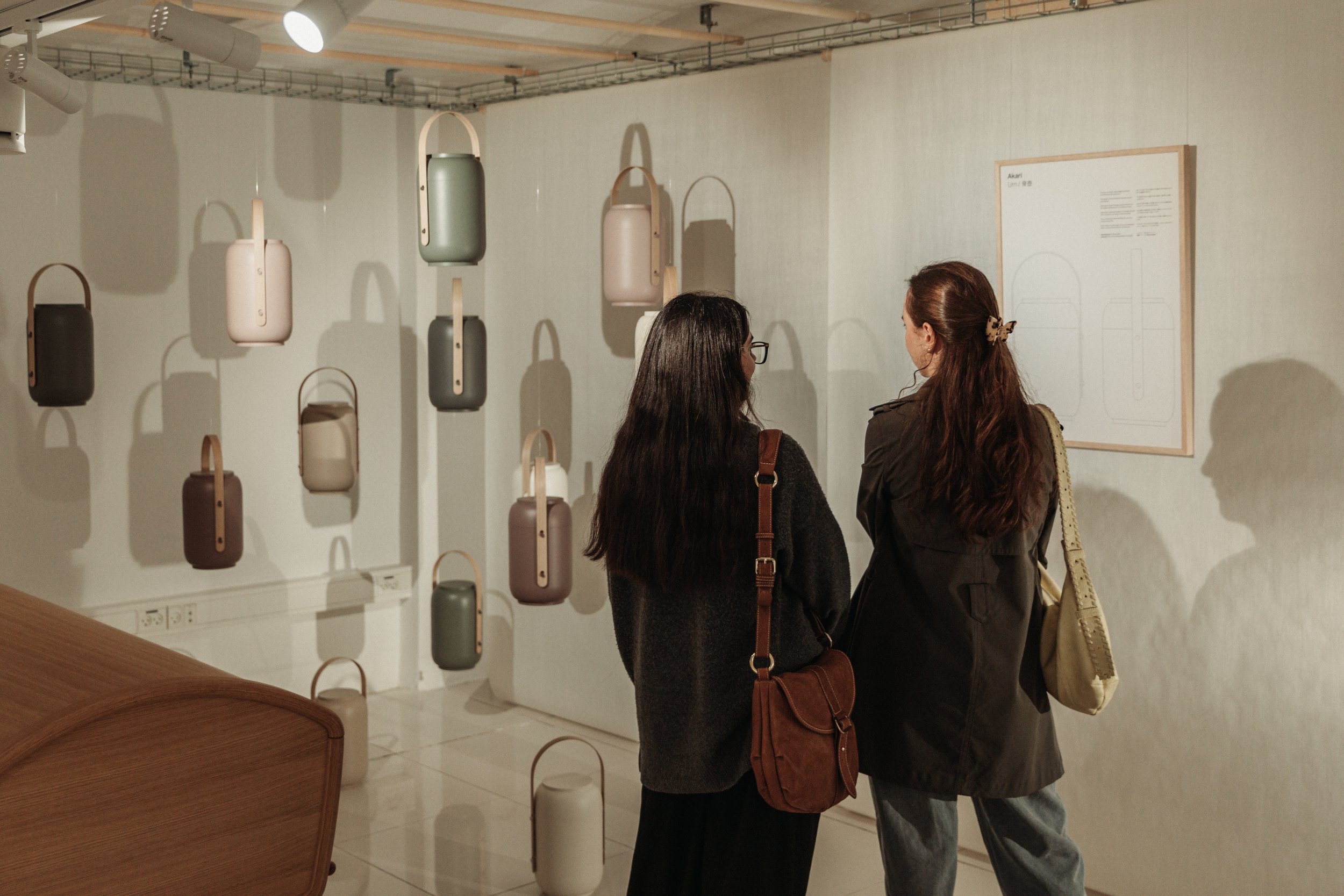 Two women with dark and reddish hair standing in an art gallery, viewing a wall display of pastel-colored handbags with wooden straps, hanging from the ceiling. The gallery has a white wall and lighting fixtures overhead.