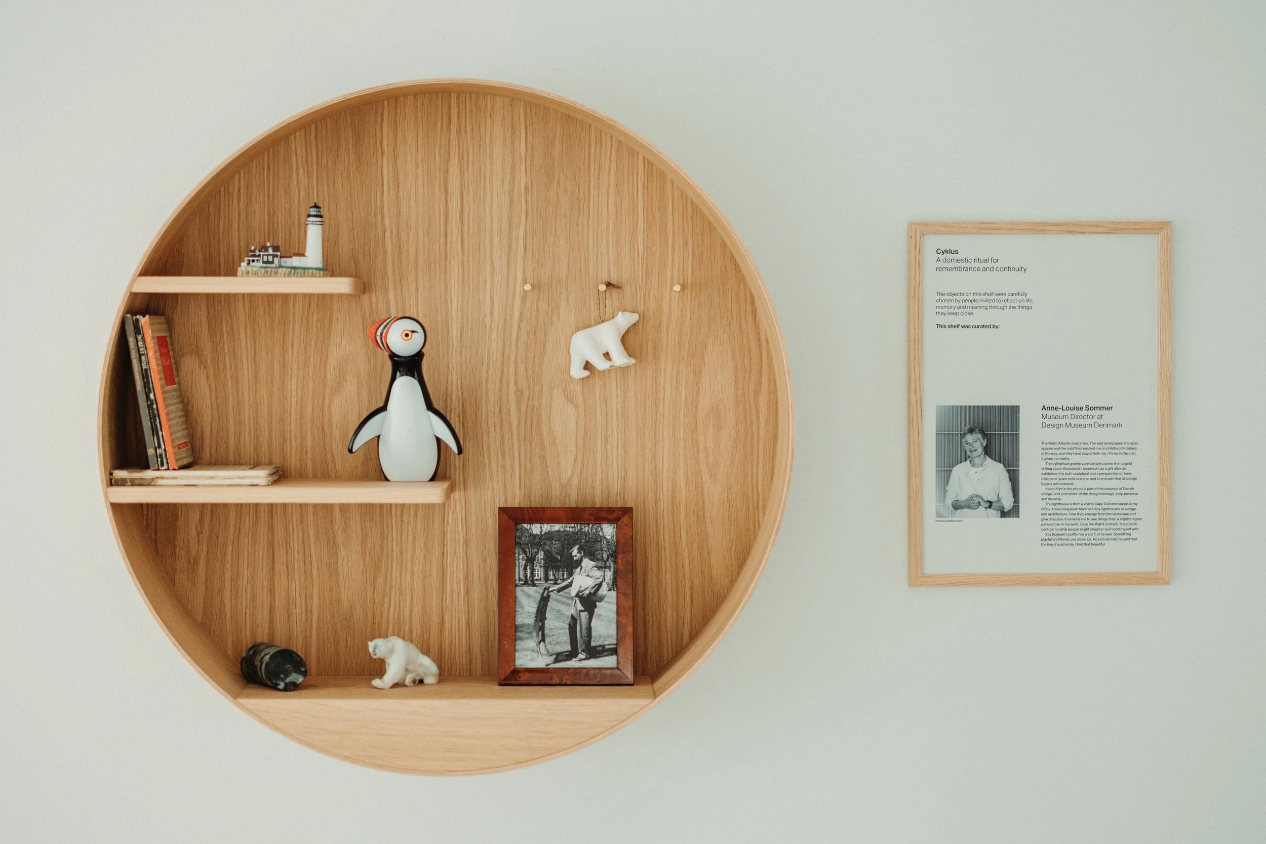Round wooden shelf with decorative items including a model lighthouse, a penguin figurine, a polar bear figurine, a black and white stone, a framed black and white photograph of a person, and a small sculpture of a polar bear. To the right of the shelf, there is a framed informational poster on a wall.