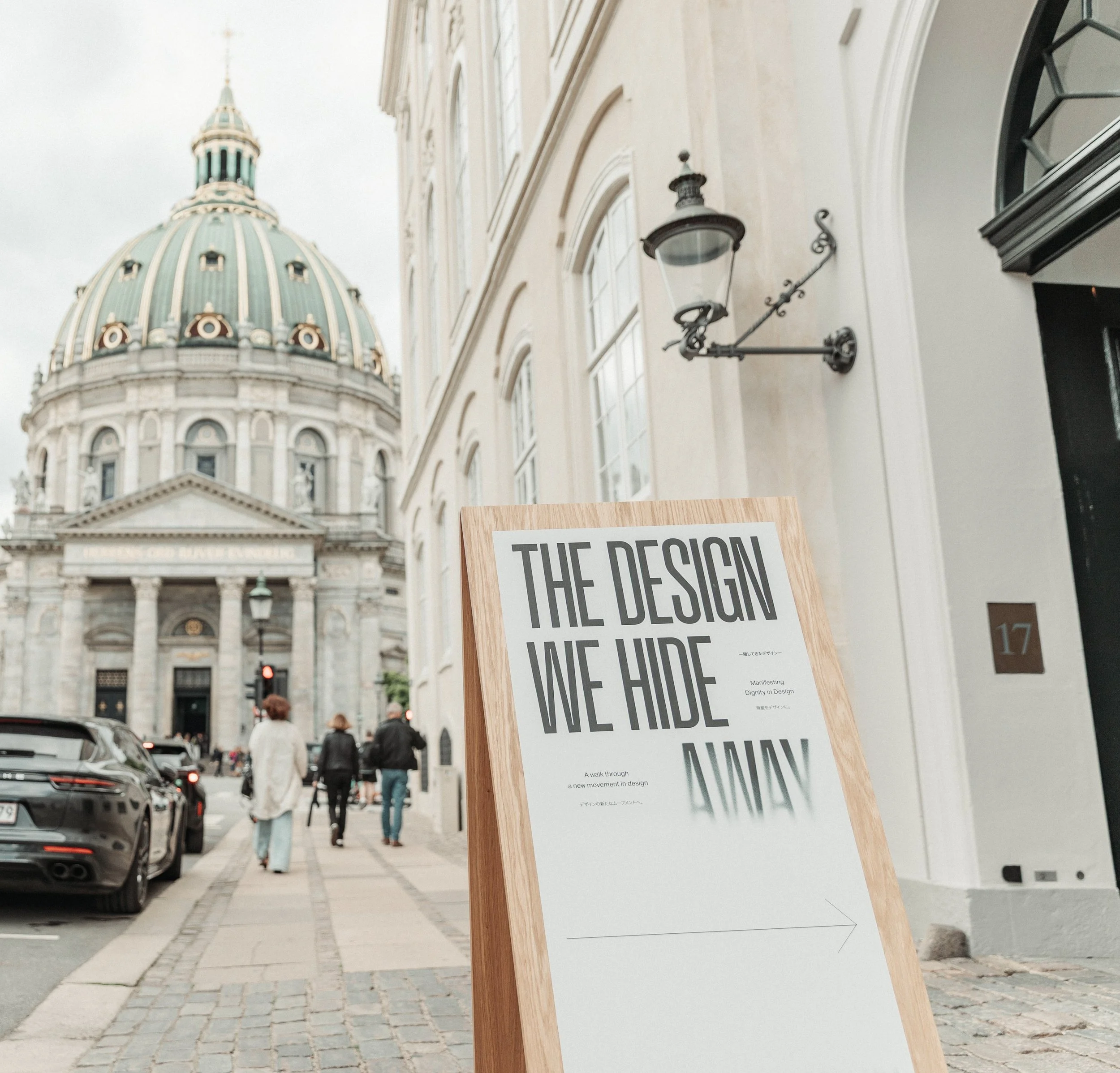 Street scene with a sign on a wooden stand reading 'THE DESIGN IN THE HIDE.' Background shows historic building with a domed roof, people walking, and parked cars.