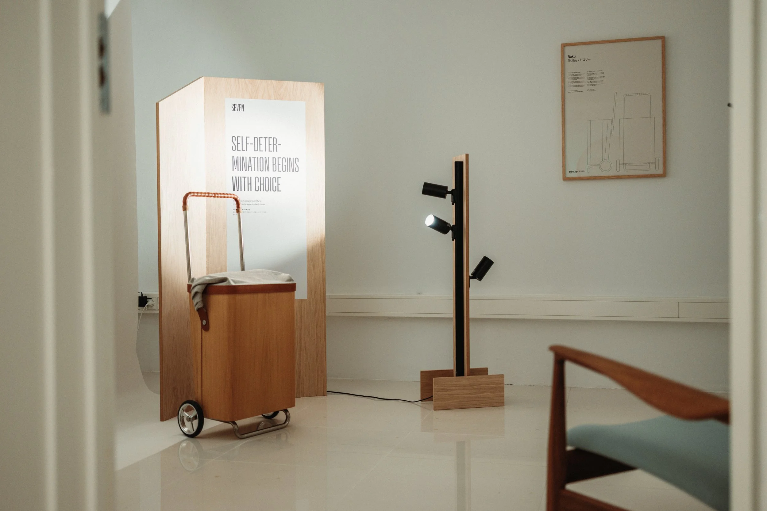 An art exhibit features a wooden sign with text and a small trolley cart, with a framed schematic drawing on the wall behind.