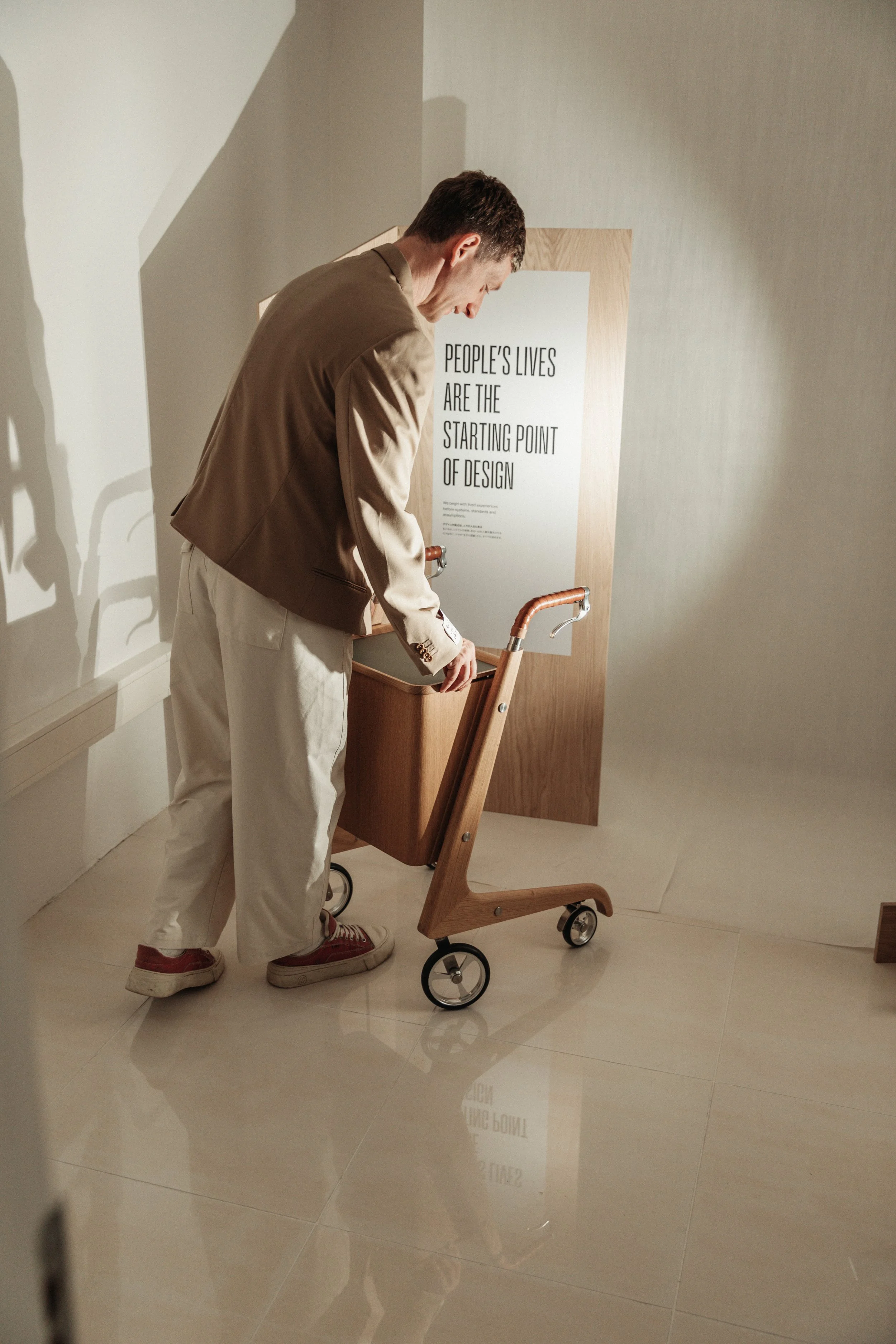 A man with dark hair wearing a beige blazer, white pants, and red sneakers, leaning over a wooden and metal cart with wheels, in front of a sign that reads 'People's Lives Are The Starting Point of Design', in an indoor setting with light-colored tiled floor and neutral walls.