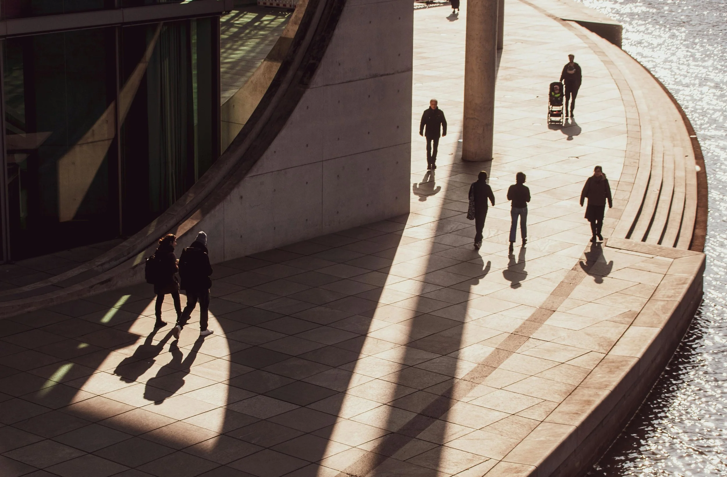 People walking on a sidewalk beside a river with reflections of sunlight, with large concrete and glass structures nearby.
