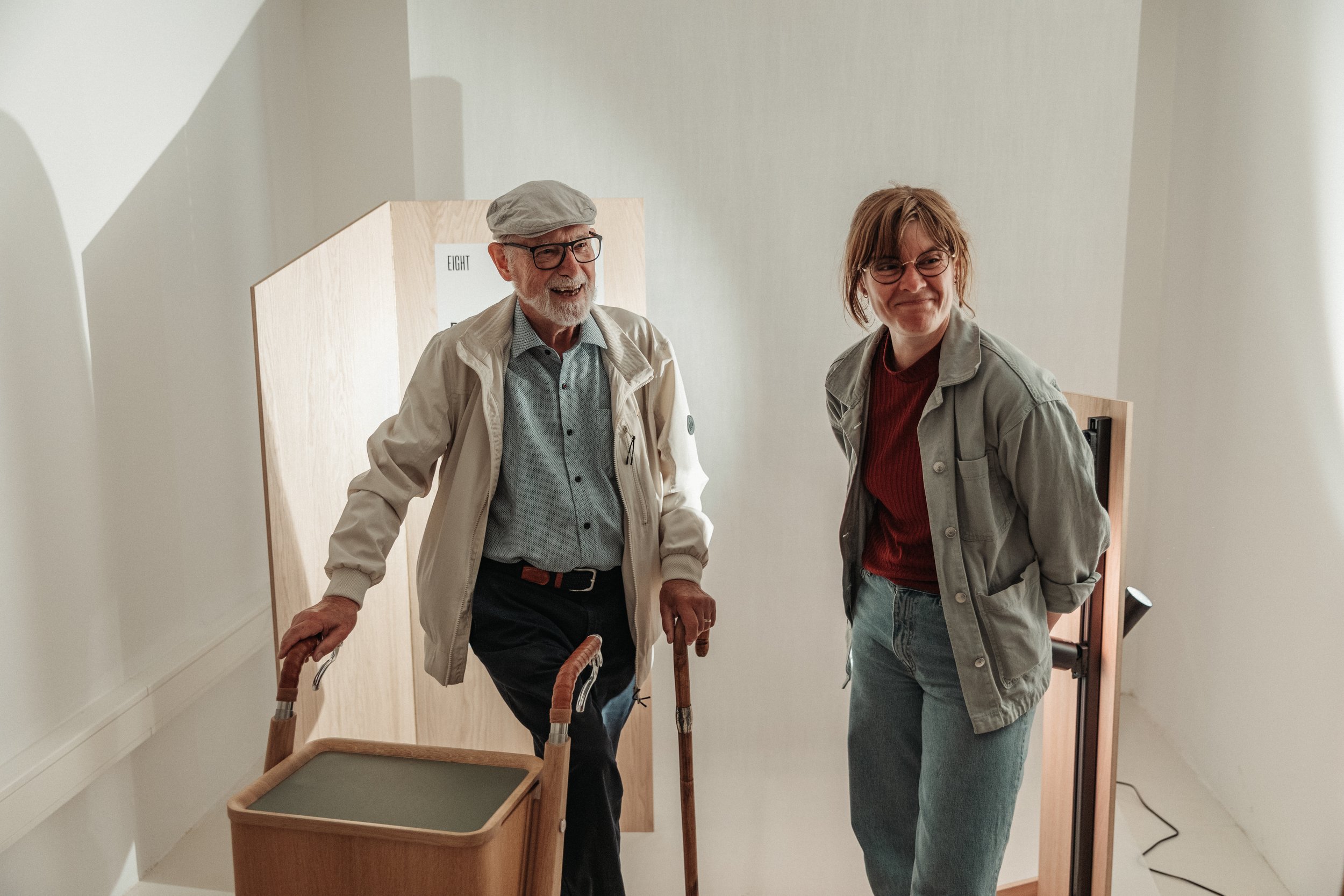 An elderly man with glasses, a gray hat, and a beige jacket, using a cane, standing next to a woman with glasses, short hair, wearing a gray jacket and a red sweater, both smiling and walking through a doorway. There is a wooden box labeled 'EIGHT' and a walker in the foreground.