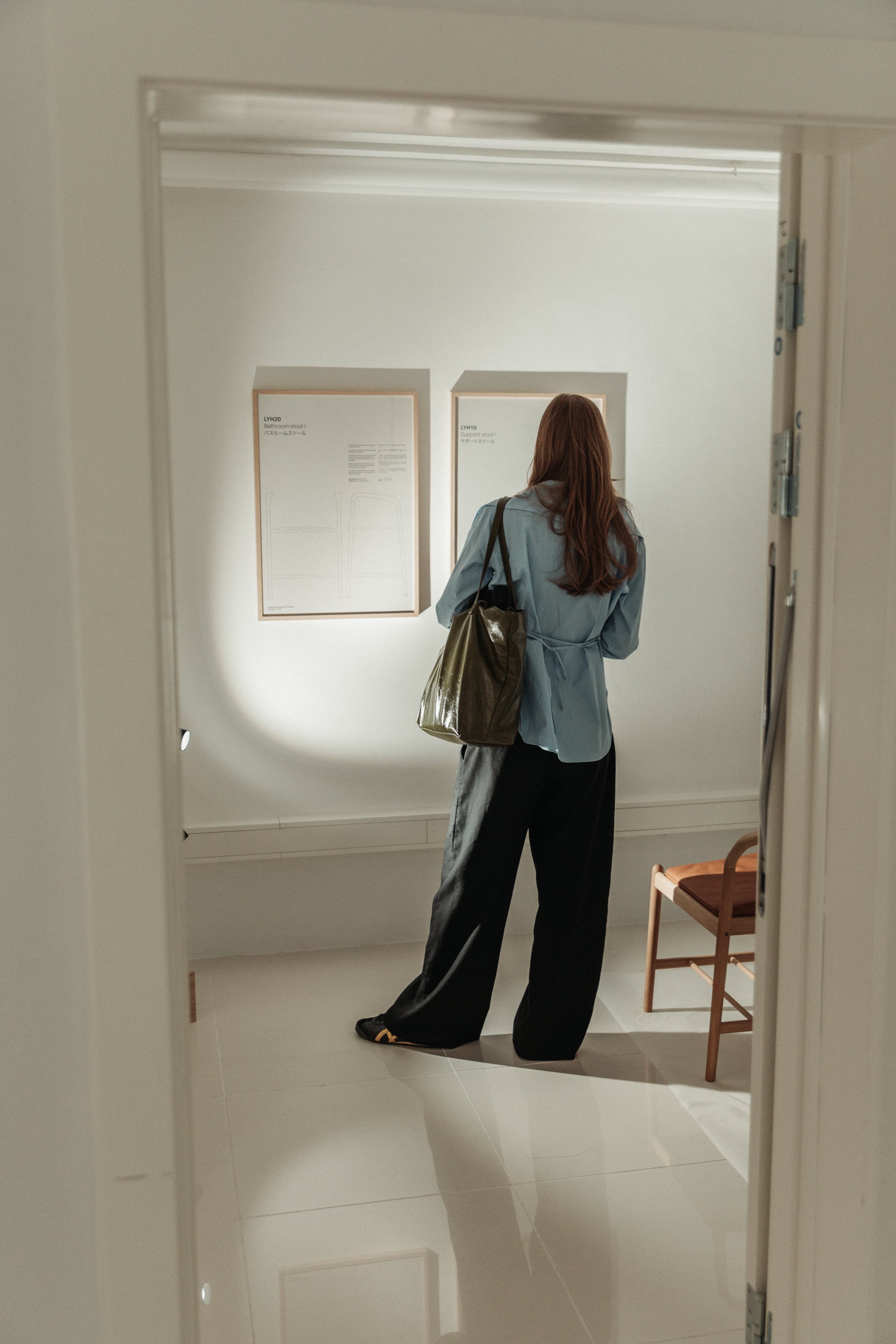 A woman with long brown hair, wearing a light blue coat and black pants, carrying a large metallic bag, seen from behind, standing in front of framed artwork on a white wall in an art gallery or museum.