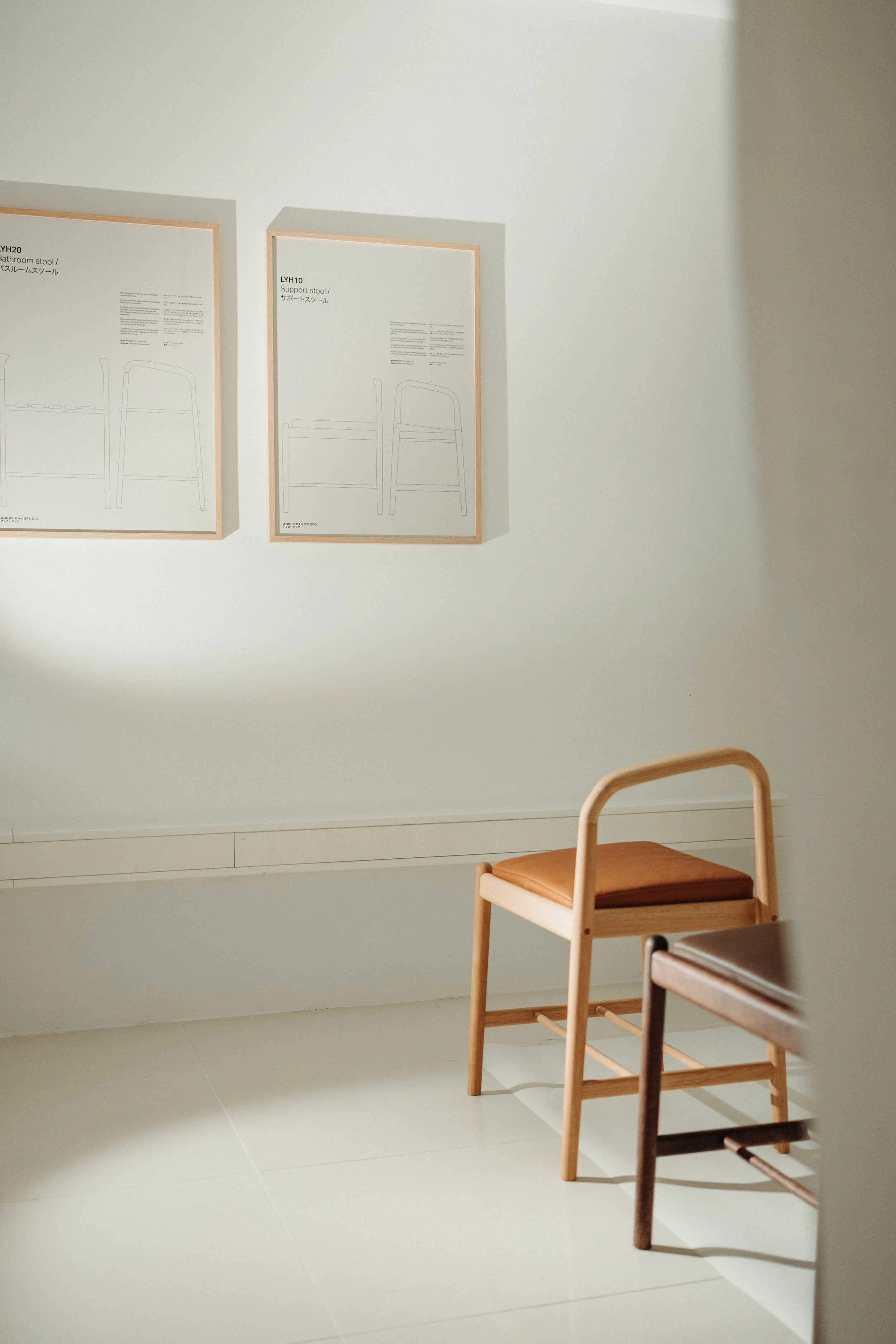 Two chairs with wooden frames and brown cushions in a light-colored room. The chairs are positioned in front of a plain white wall with two framed diagrams of furniture labeled 'Support stool' and 'Bathroom stool'.