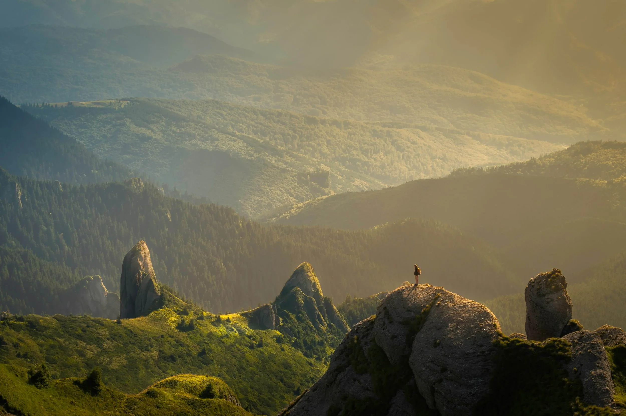 A person standing on a rocky outcrop overlooking a lush green mountainous landscape with ridges and valleys during sunrise or sunset.