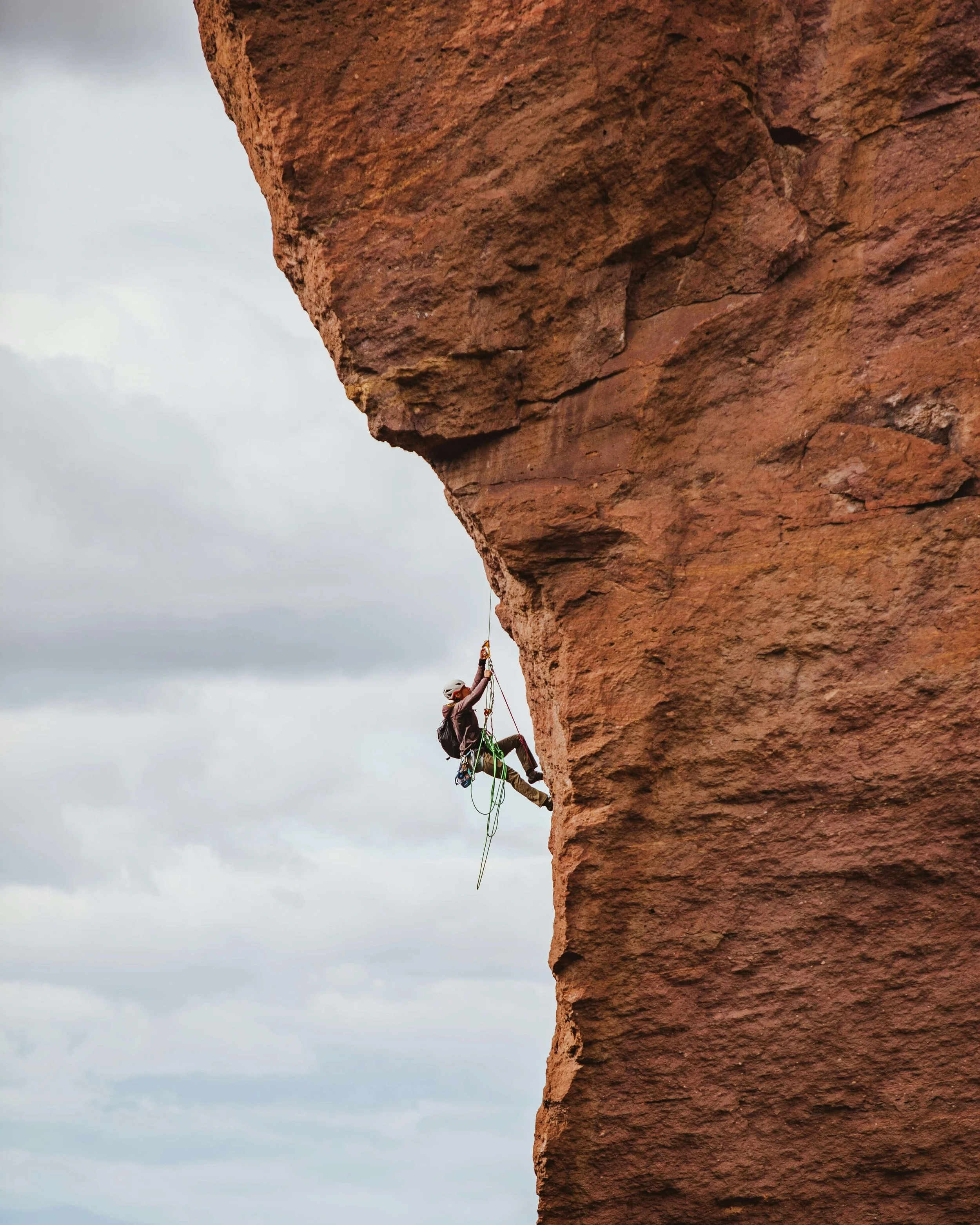 A person wearing a helmet and climbing harness is rock climbing on a large red rock formation with a cloudy sky in the background.