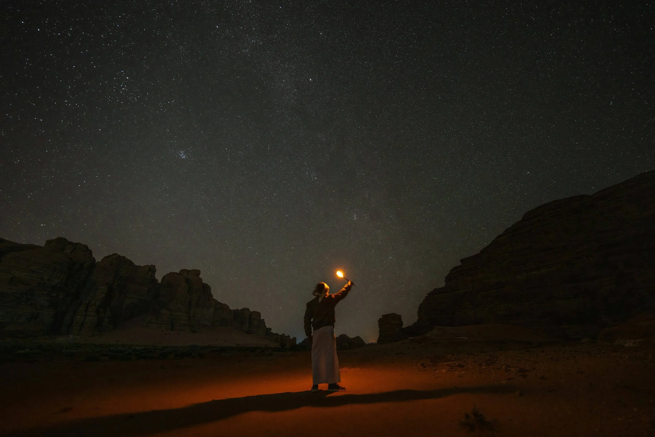 Person standing outdoors at night in a canyon, holding a lit torch under a starry sky.