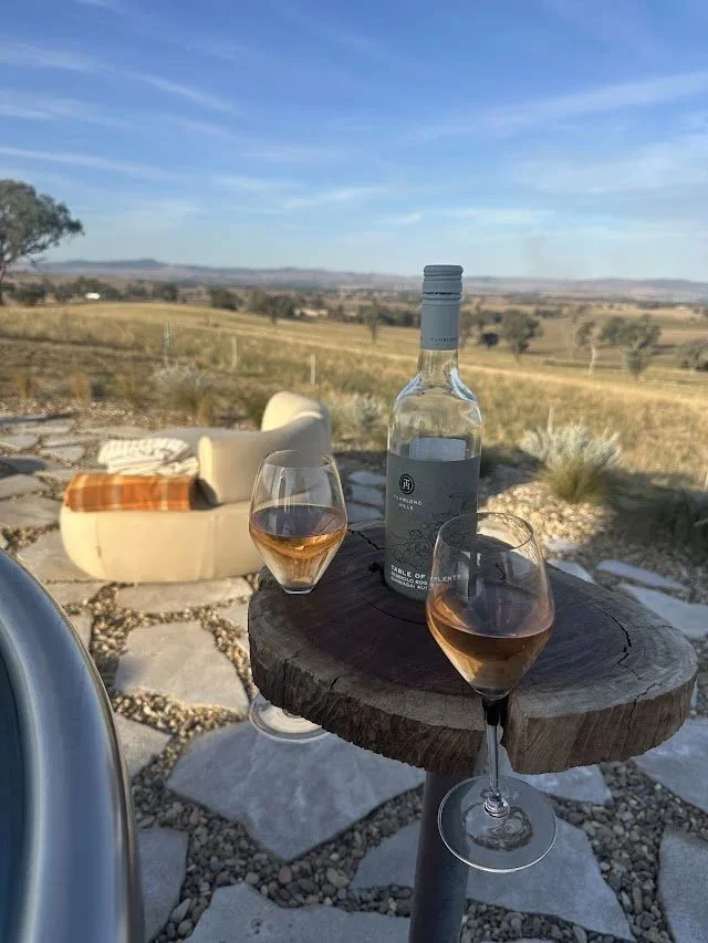 Two wine glasses filled with rosé wine on a small round wooden table outdoors, with a bottle of wine, a curved white chair with an orange and brown pillow, and a scenic landscape of fields and hills in the background under a blue sky.