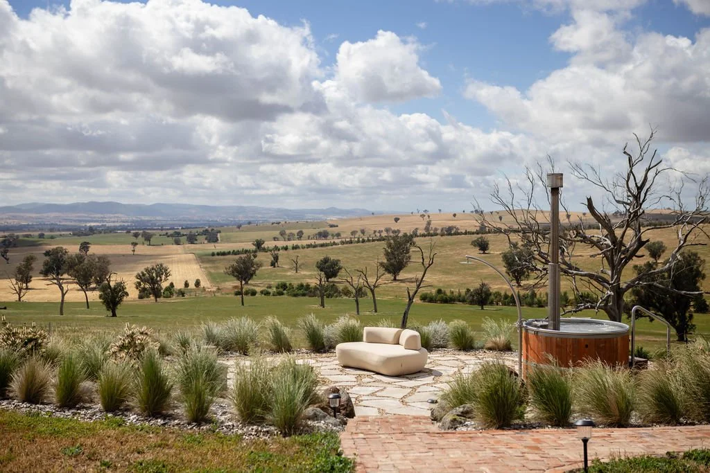 Outdoor patio with a beige lounge chair, surrounded by green bushes and tall grasses, overlooking a scenic rural landscape with rolling hills, scattered trees, and a cloudy sky.