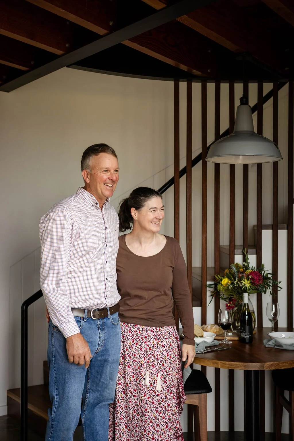 A man and woman smiling together in a dining area with a round wooden table set for a meal, a bottle of wine, wine glasses, bread, and a flower arrangement. The background features wooden slats and a hanging pendant light.