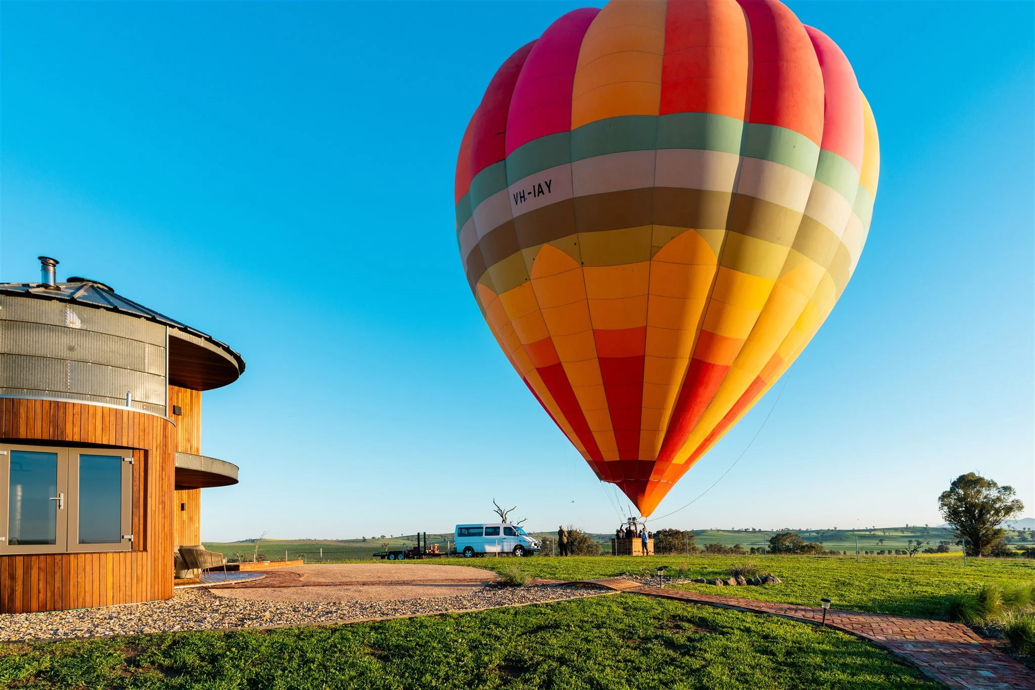 Colorful hot air balloon floating above a grassy field near a modern wooden building with a curved rooftop during daytime.