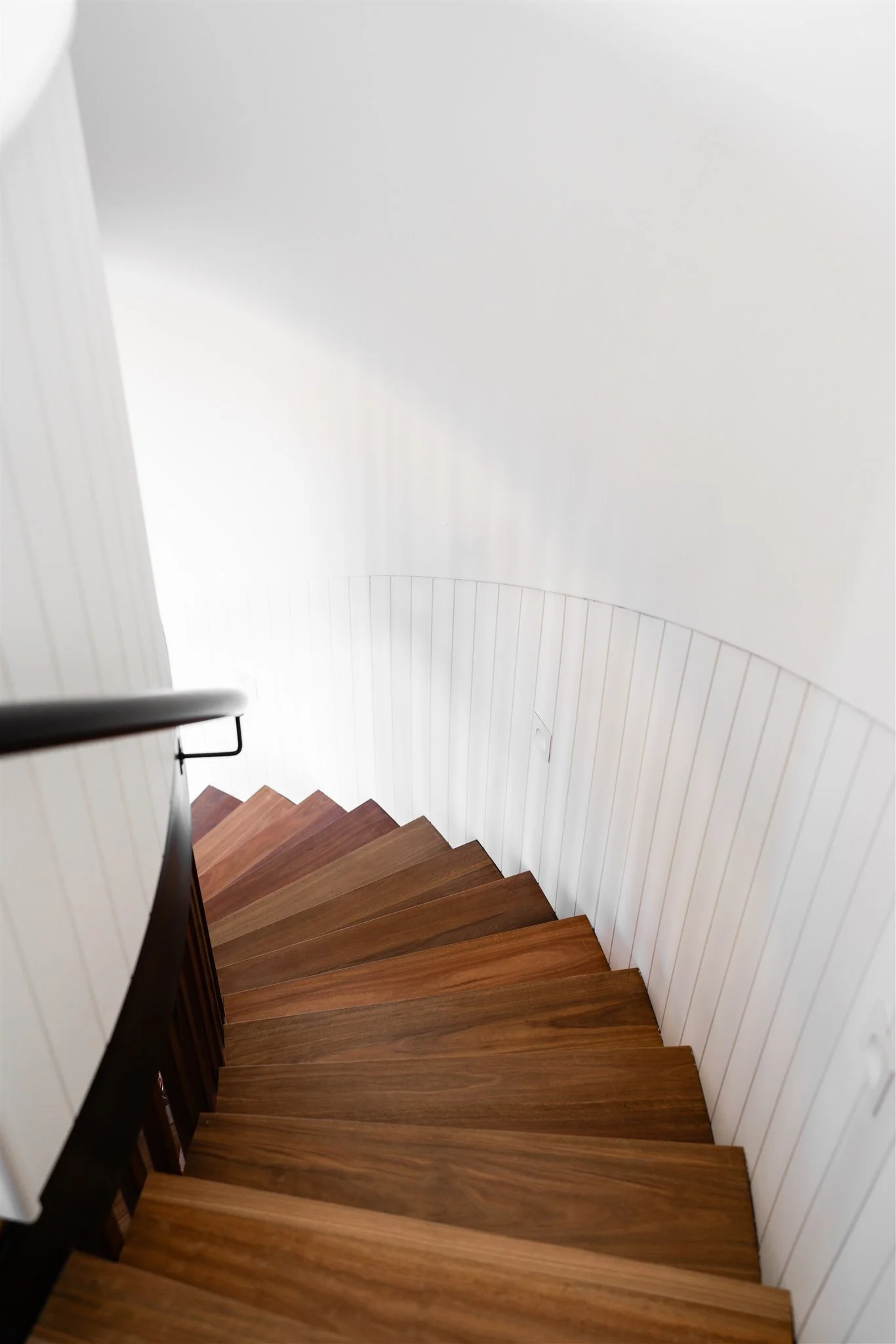 View of a staircase with wooden steps and a dark wooden handrail, winding down along a white wall.
