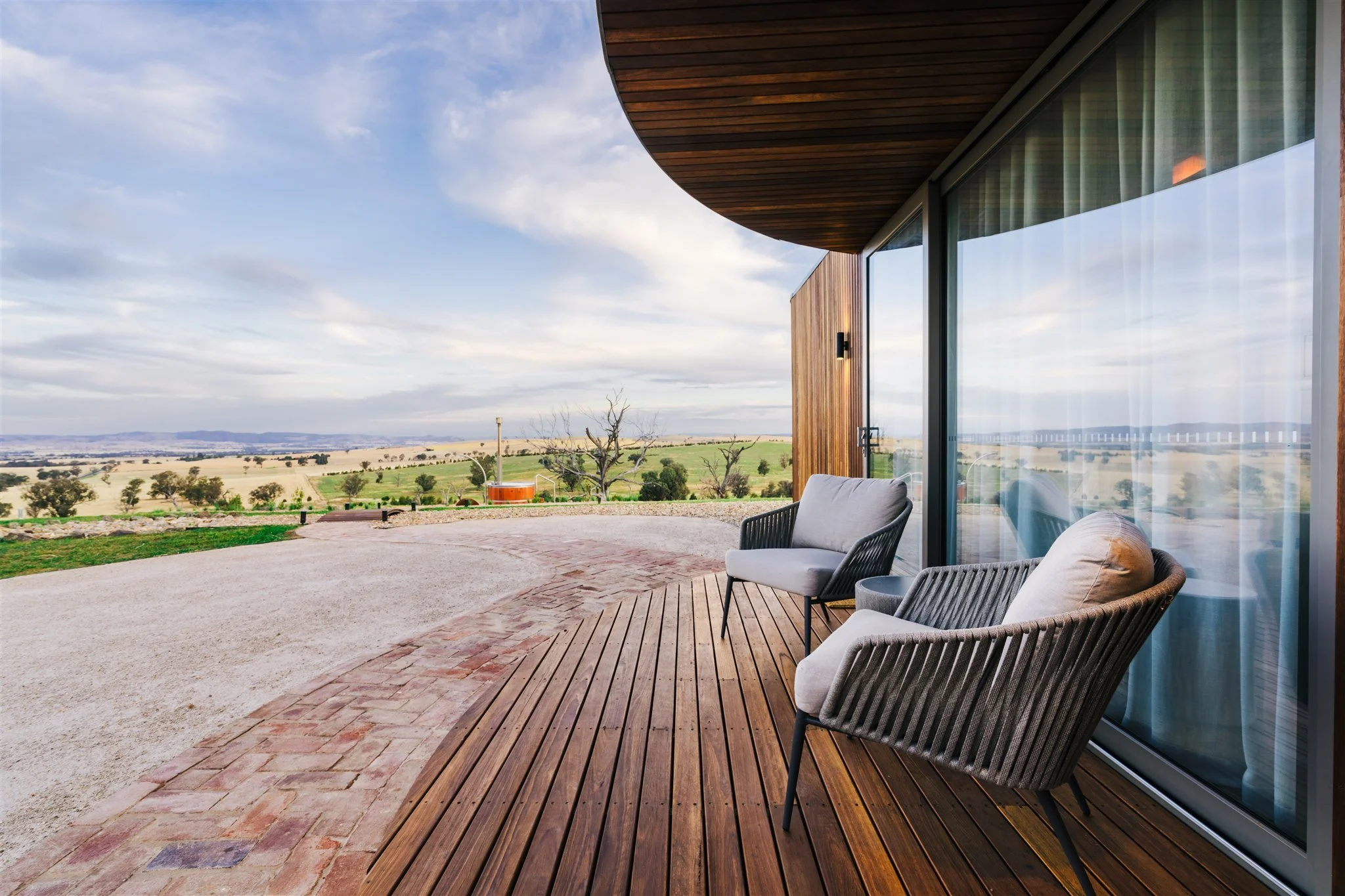 Modern house exterior with a curved wooden overhang, large glass sliding doors, outdoor seating with white cushioned chairs on a wooden deck, and a scenic view of rolling hills and fields under a partly cloudy sky.