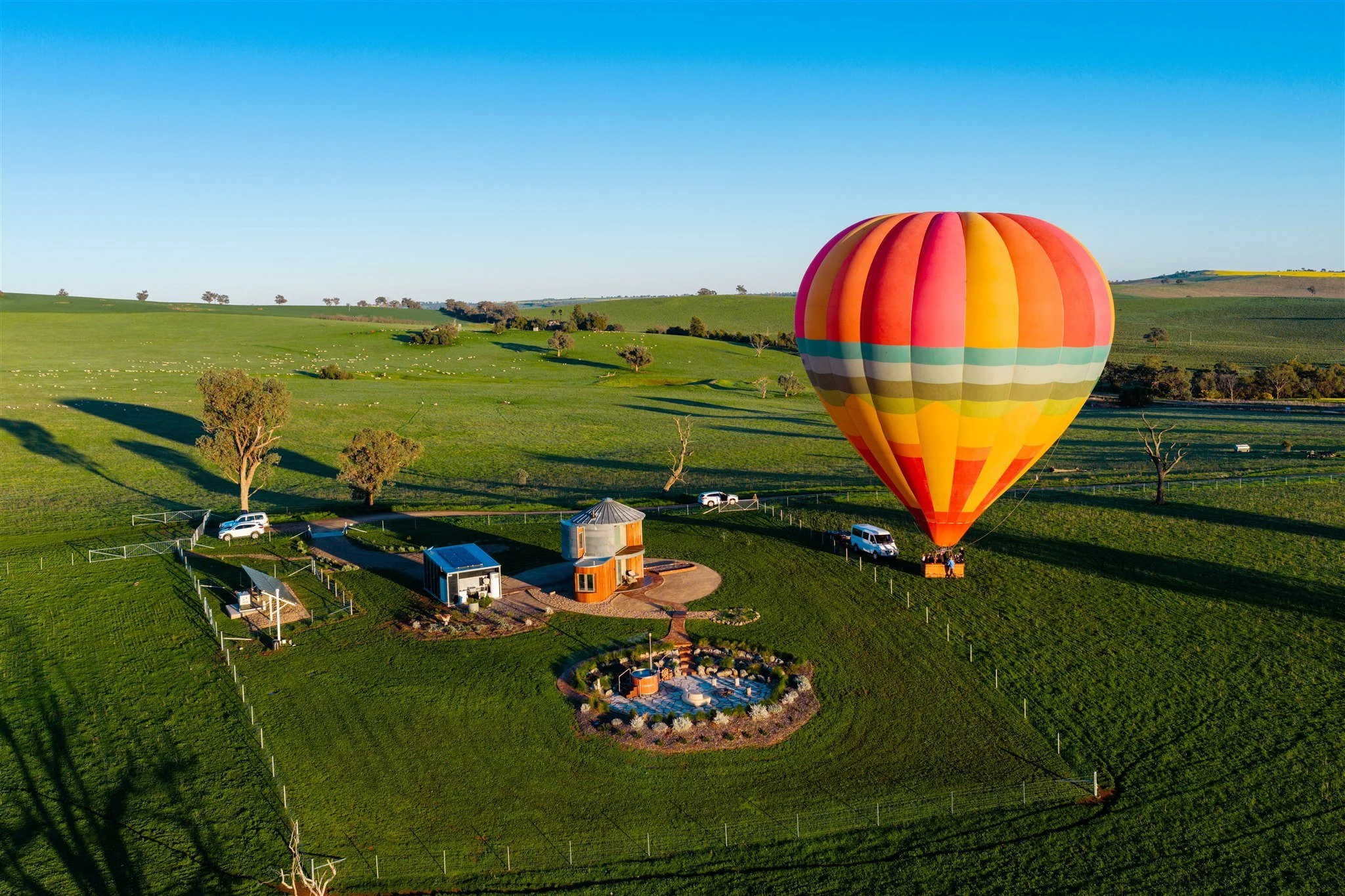 A colorful hot air balloon inflates on a green field with rolling hills, trees, and a clear blue sky in the background.