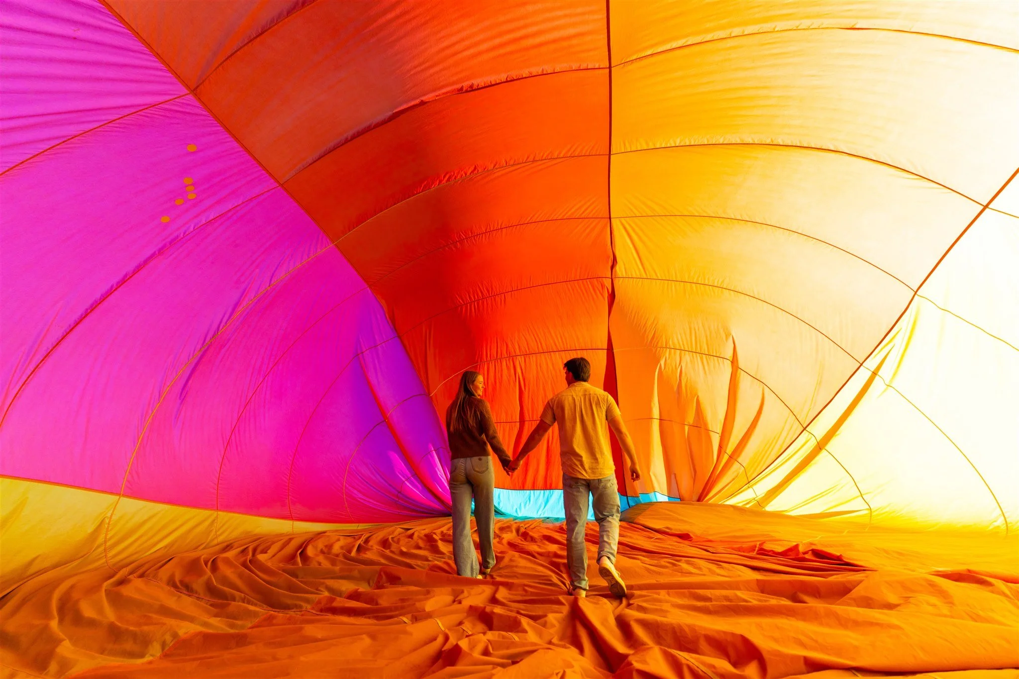 Two people holding hands inside a hot air balloon envelope with vibrant pink, orange, and yellow panels.