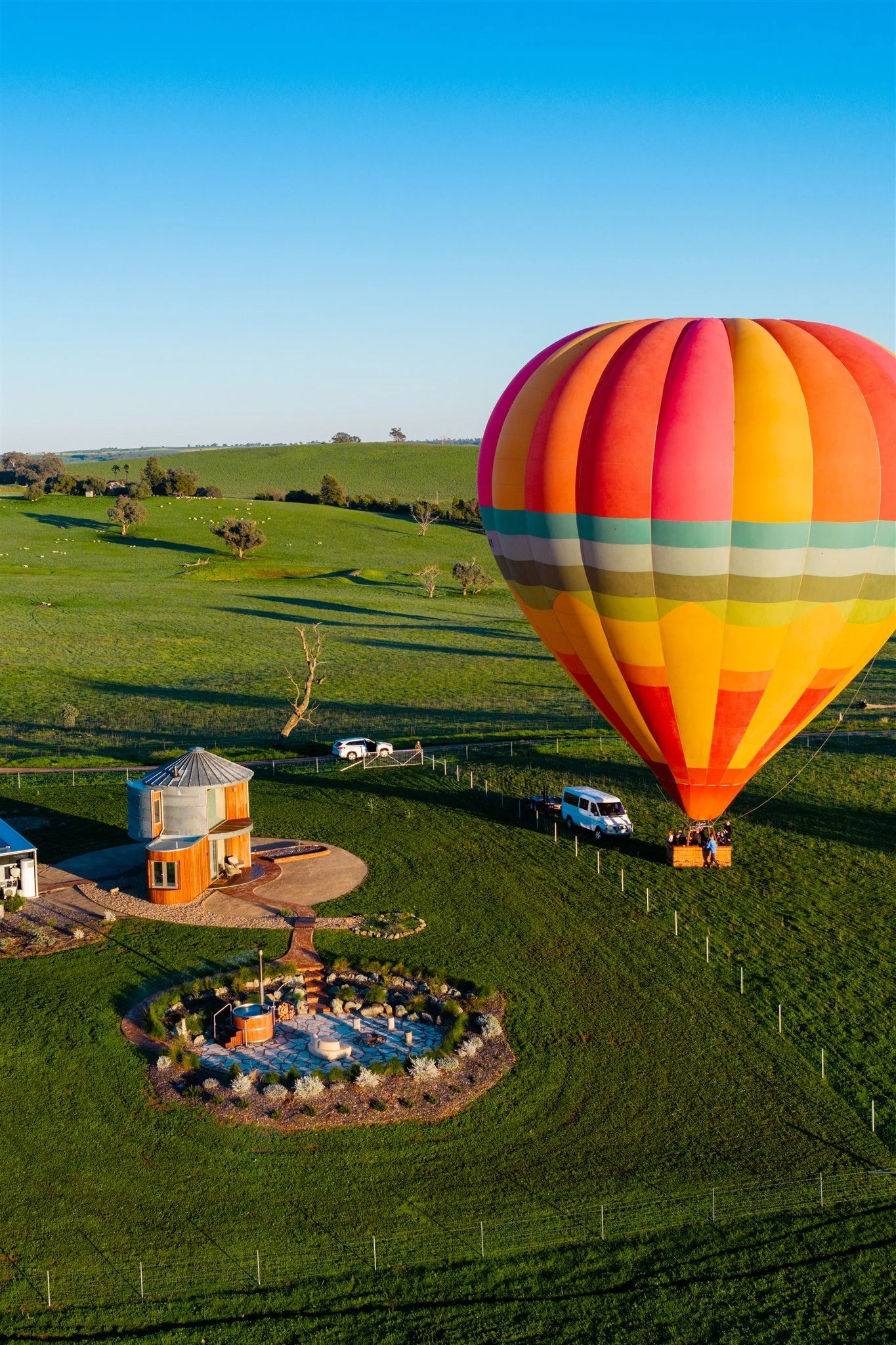 A colorful hot air balloon floating over a lush green landscape with rolling hills, trees, and small buildings, under a clear blue sky.