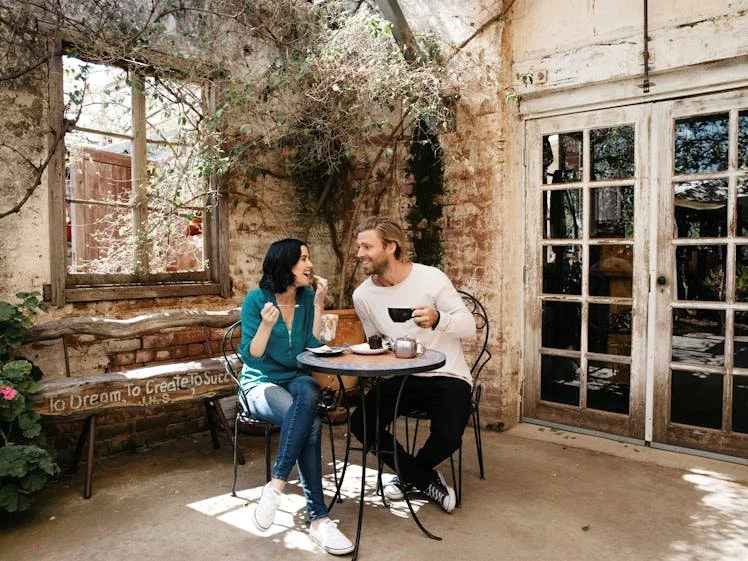 A man and woman sit at a small round table outside a rustic building, enjoying coffee and conversation with sunlight and hanging plants around them.