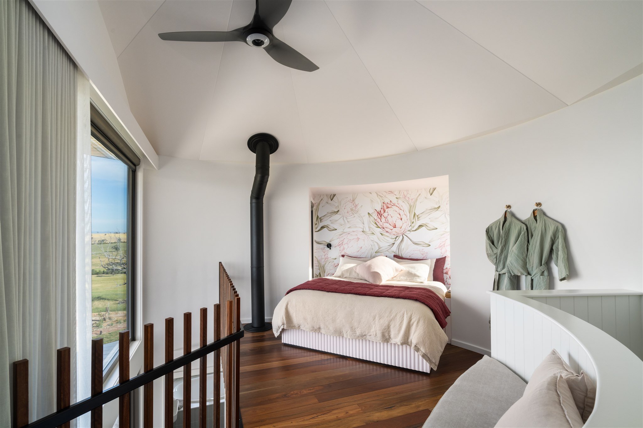 A cozy bedroom with a bed, floral headboard, and sunlight streaming through large windows, featuring wooden floors, a ceiling fan, and a black pipe ceiling vent.