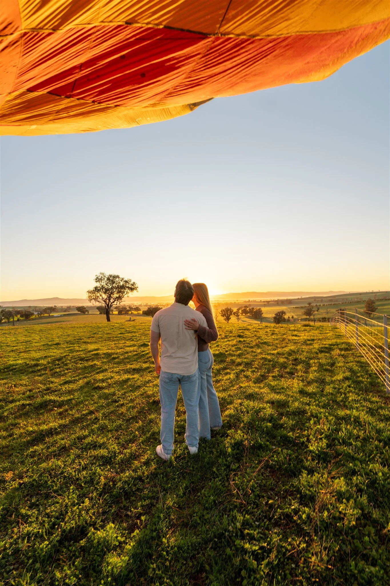 A couple embracing and looking at the sunset in an open grassy field with a few trees and hills in the background, viewed from behind with a large decorative orange and yellow umbrella overhead.