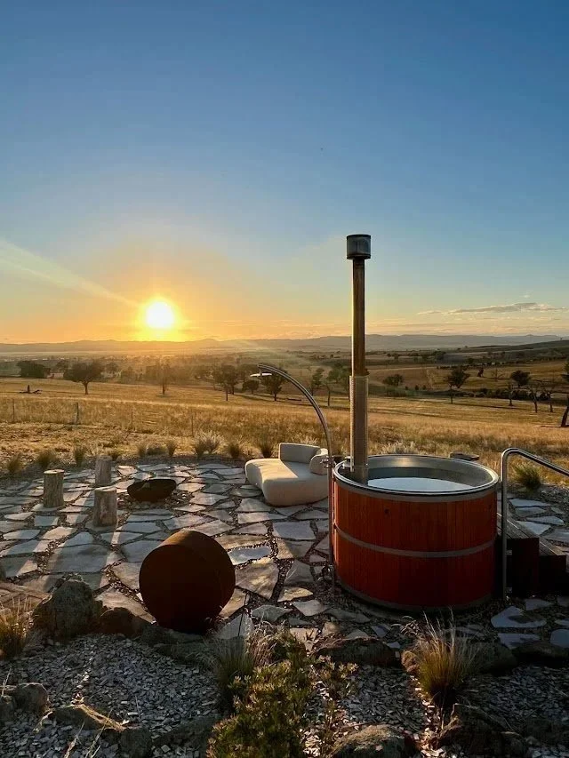 Outdoor hot tub with wooden exterior on a stone patio, set in a rural landscape with open fields, trees, and a sunset sky.