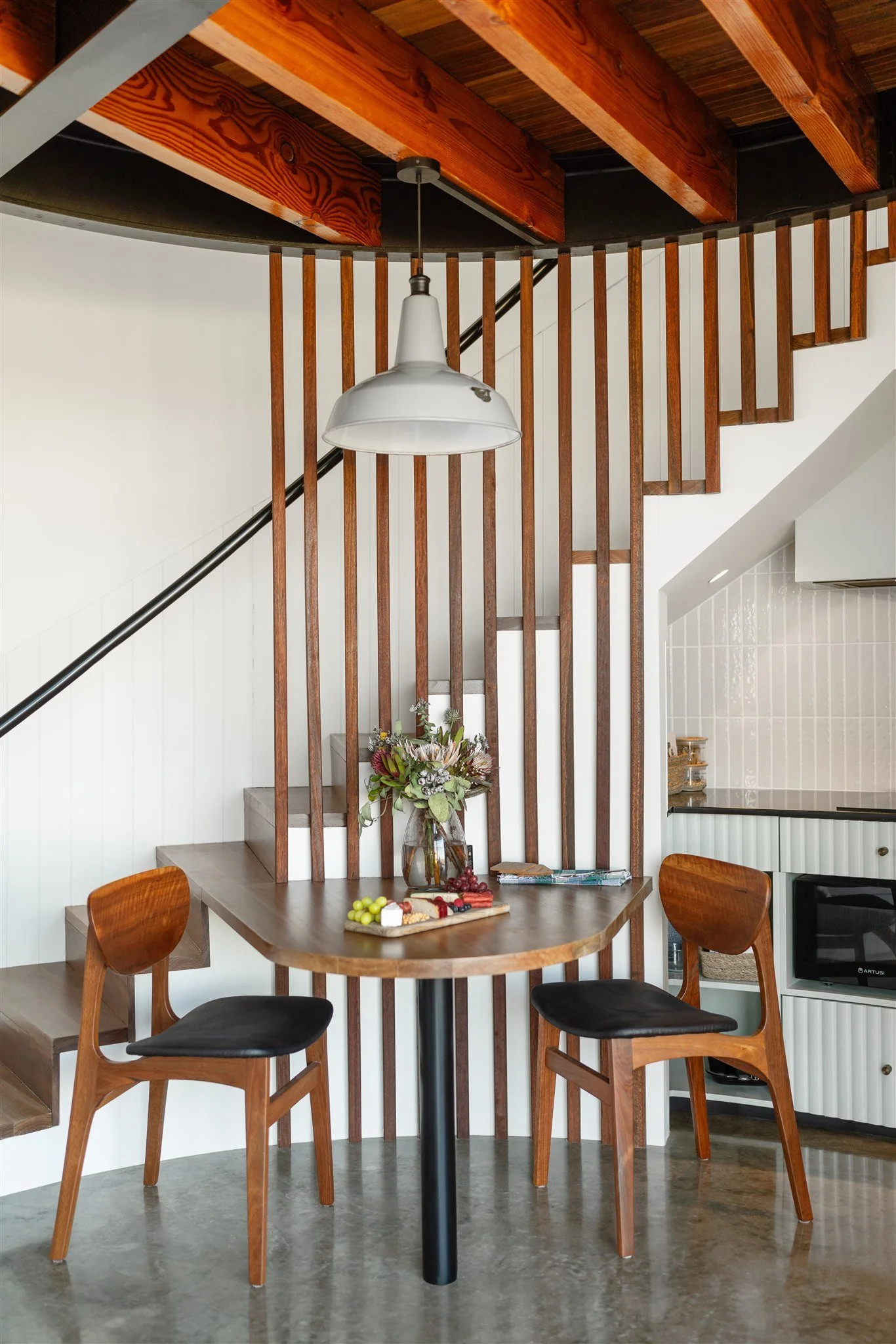 Wooden dining table with four chairs, decorated with fruit, cheese, and a flower arrangement; background features a staircase with vertical wooden slats and a ceiling pendant light.