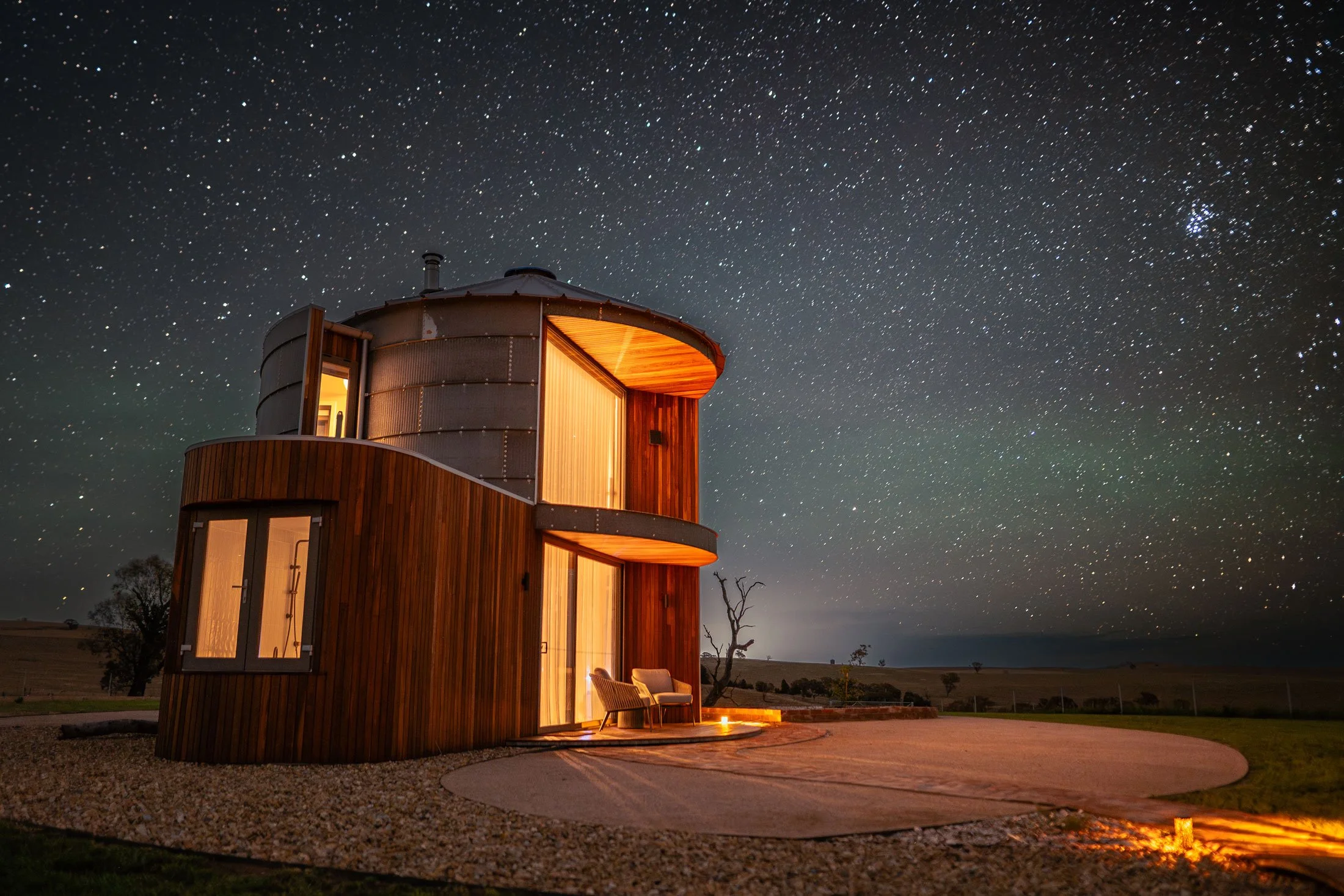 A modern, curved wooden house with large windows, illuminated from inside, set against a starry night sky. There are outdoor chairs and a gravel path in front of the house.
