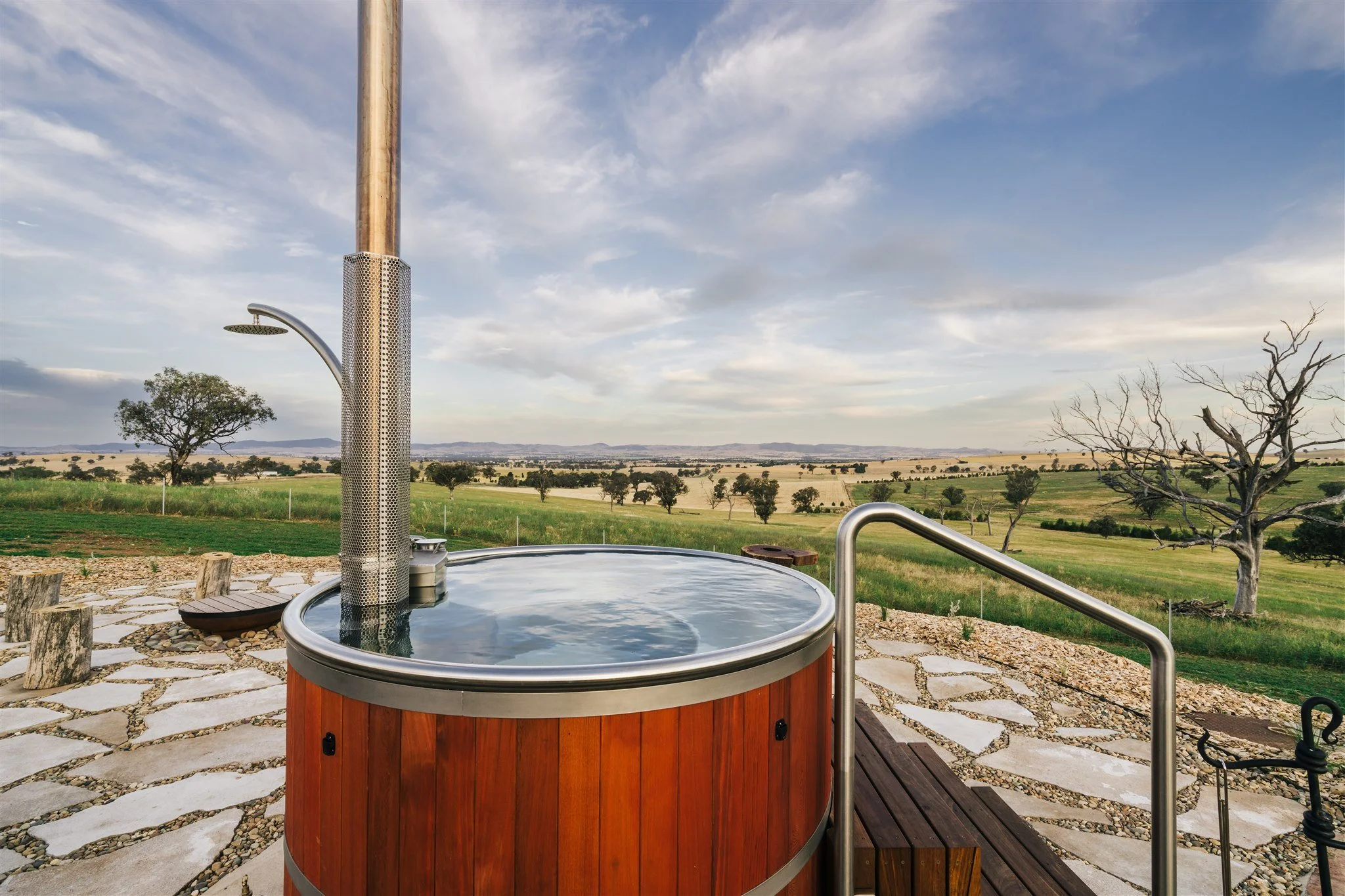 An outdoor hot tub with a wooden exterior on a patio with a stone path, overlooking a rural landscape with fields, trees, and a cloudy sky.