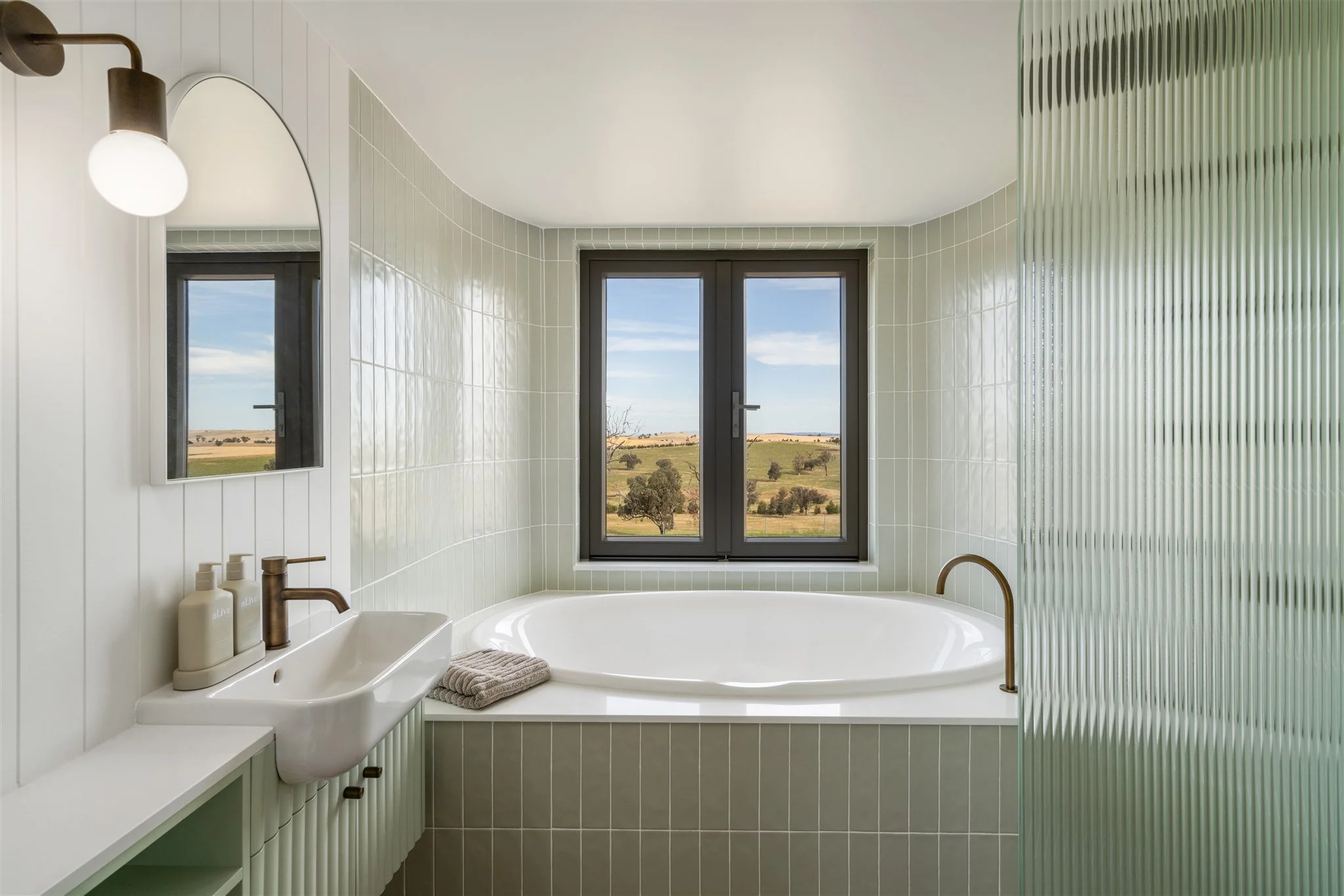 A modern bathroom with a large window offering a view of a rural landscape, a bathtub, a sink with a bronze faucet, and a frosted glass partition.