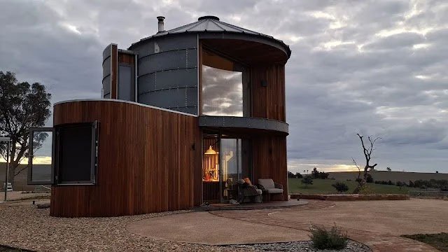 Modern house with a curved wooden exterior, large windows, and a metal upper level, situated in a rural landscape with cloudy skies.