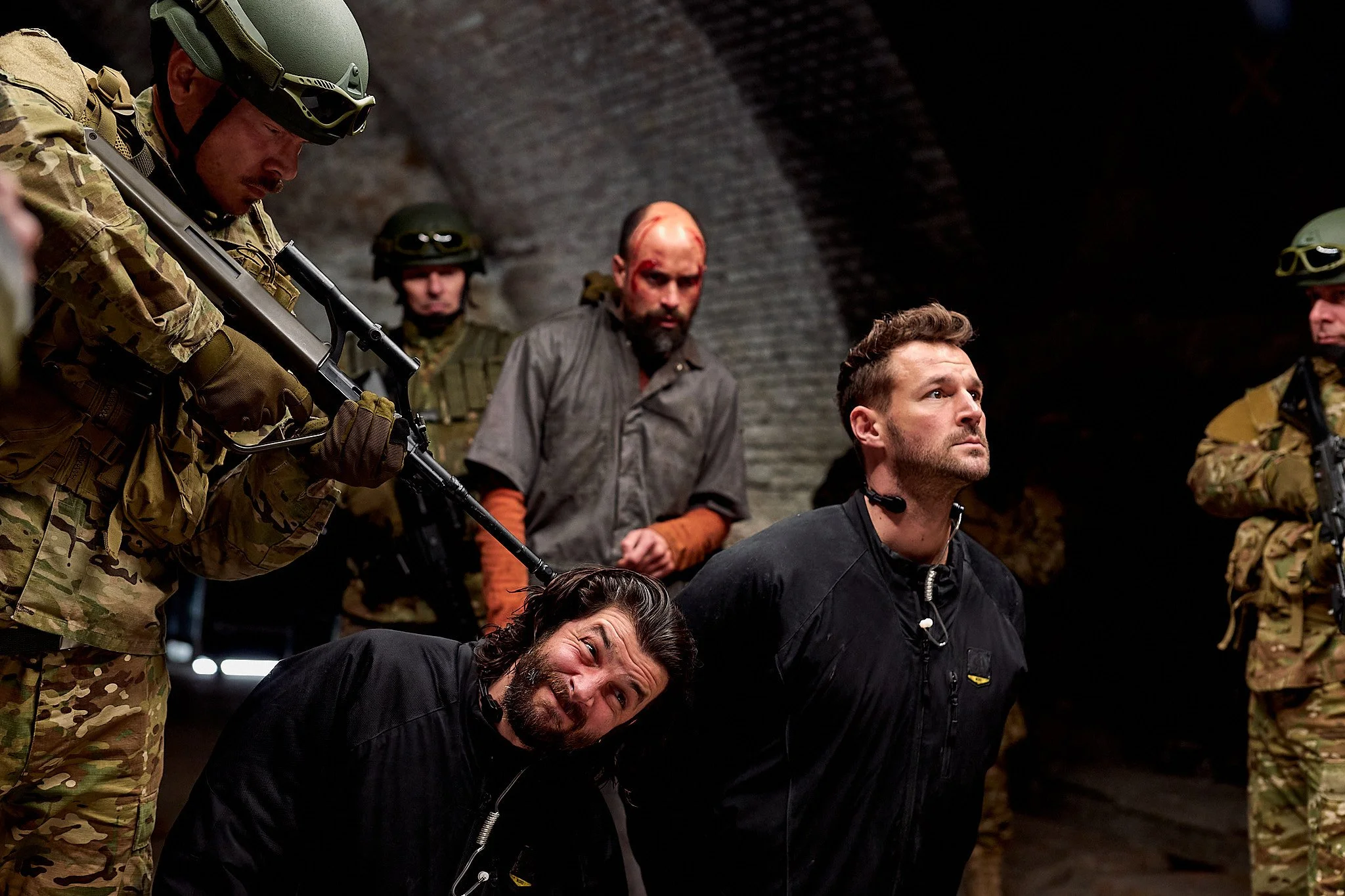 Military personnel in camouflage uniforms and helmets surrounding a man with a beard and makeup resembling a bloody head wound, and others with serious expressions is in an underground tunnel.
