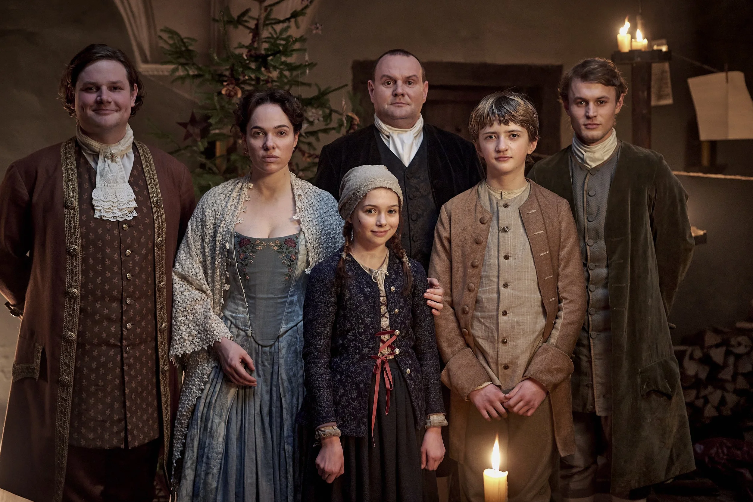 Group of six people dressed in historical clothing standing in front of a Christmas tree and candles.