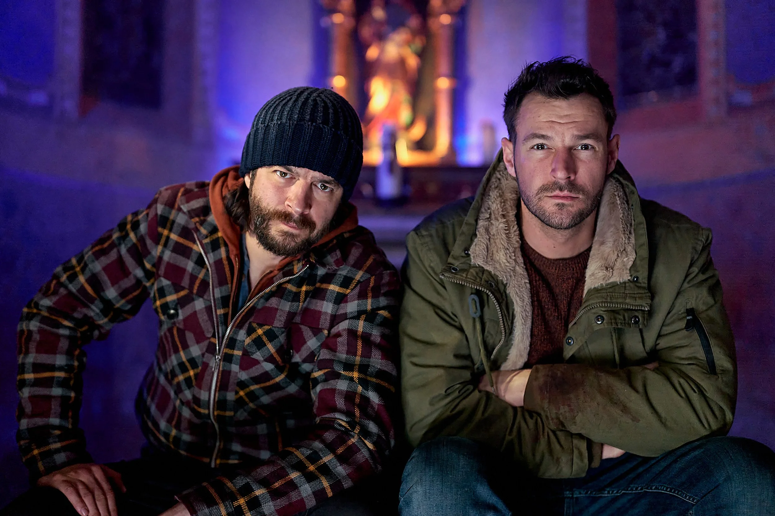 Two men sitting closely together in a dimly lit room with blue and purple lighting, with a blurred religious statue in the background. One man wears a dark knit beanie and a plaid jacket, the other has short dark hair and an olive green jacket.