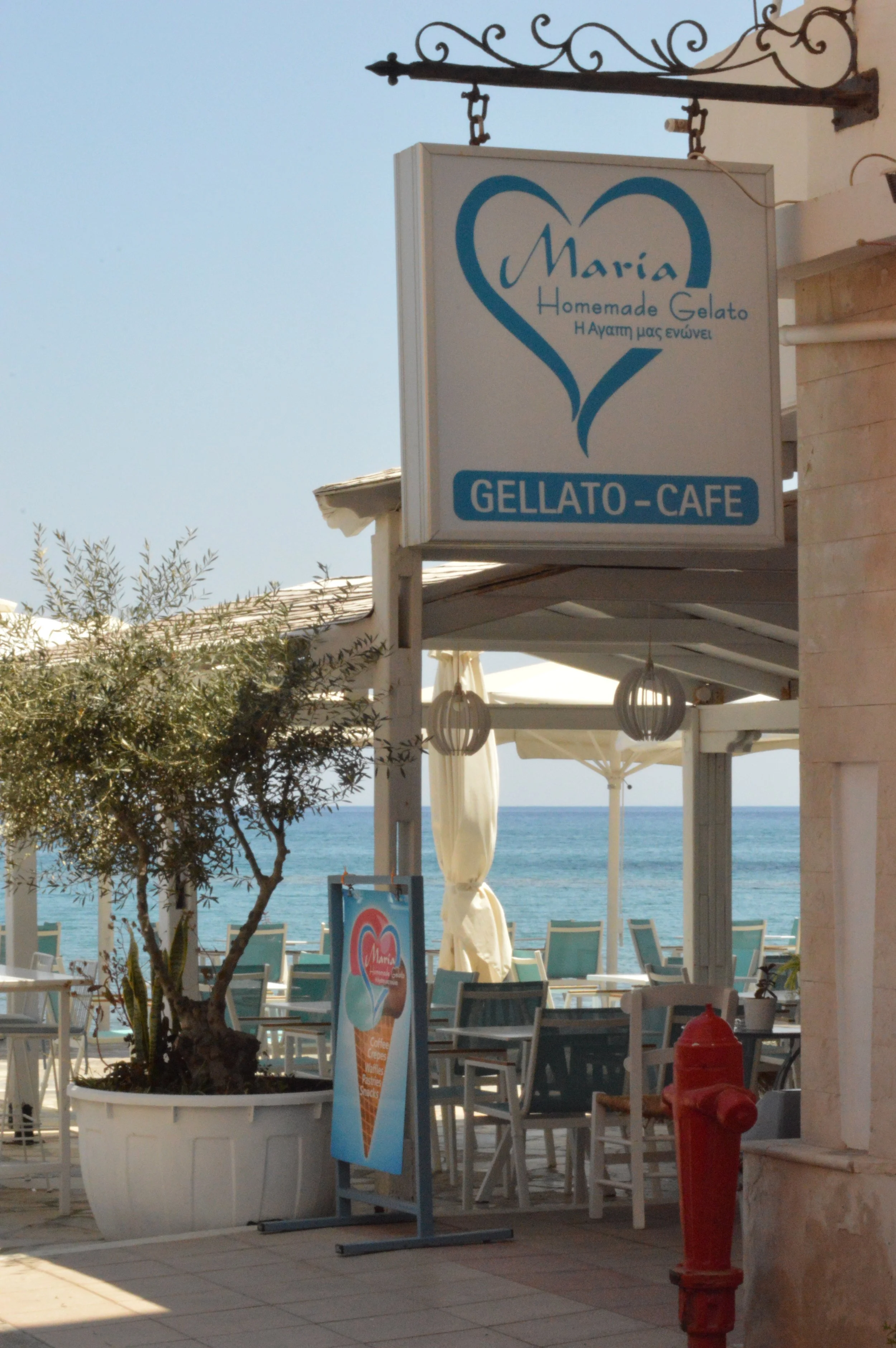 Gelateria Maria - A sign for Maria Homemade Gelato Cafe hangs above an outdoor seating area with the beach and ocean in the background. The sign features a blue heart shape and text in both English and Greek.