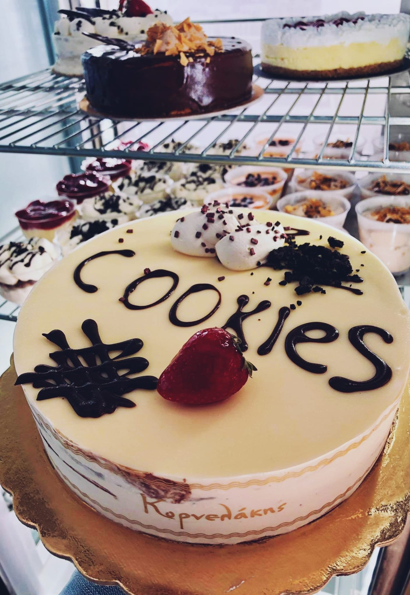 Gelateria Maria - A decorated birthday cake with strawberry, whipped cream, and chocolate writing that says 'Cookies,' with additional desserts and cakes in the background.