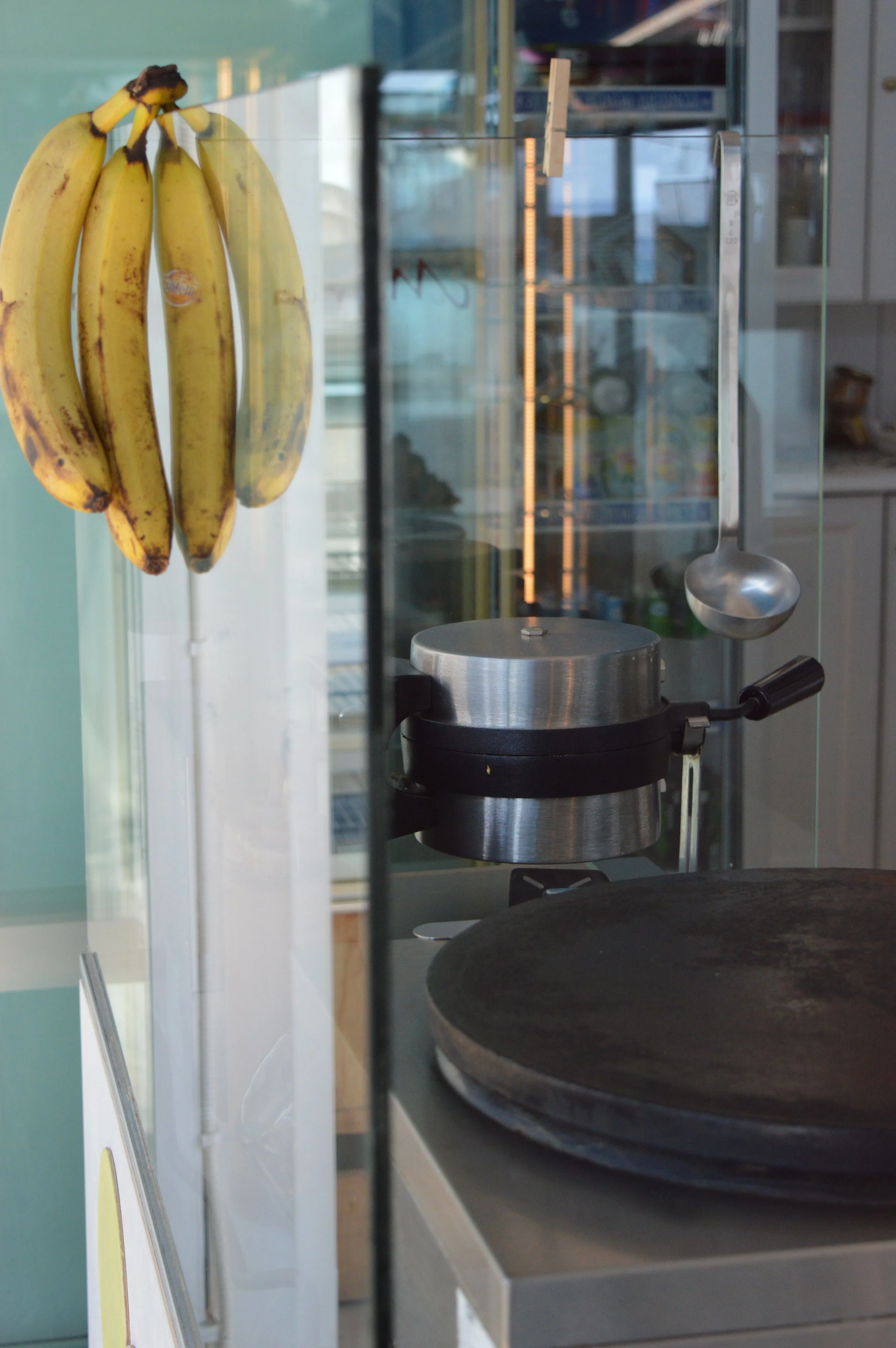 A bunch of ripe bananas hanging in a display case next to a metal scoop, a stovetop, and a glass window in a kitchen or bakery setting.