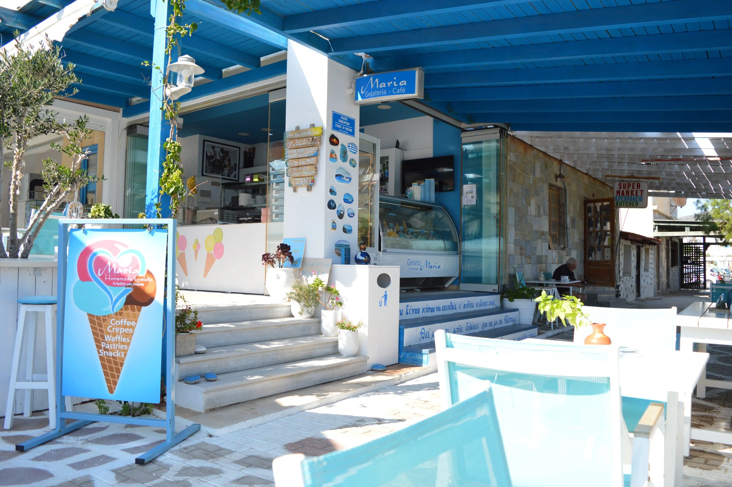 Exterior view of a small ice cream shop named Maria Gelateria Cafe, with a menu board and ice cream display case, outdoor seating with white chairs and blue cushions, potted plants, and signs advertising coffee, crepes, waffles, pastries, and snacks,