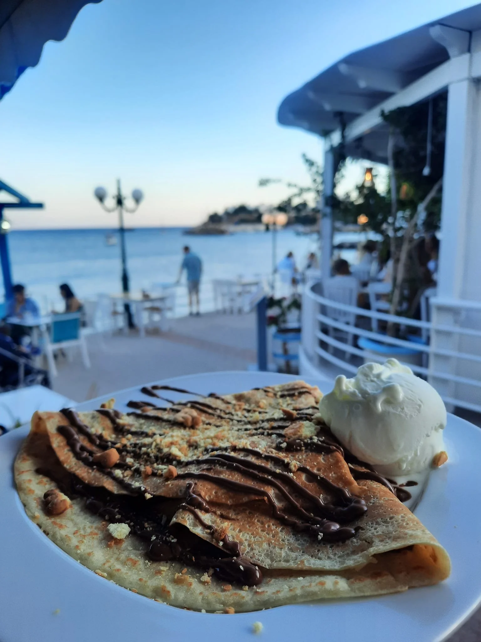 Crepe topped with chocolate sauce and crushed nuts, served with a scoop of vanilla ice cream, on a white plate with outdoor seaside dining in the background