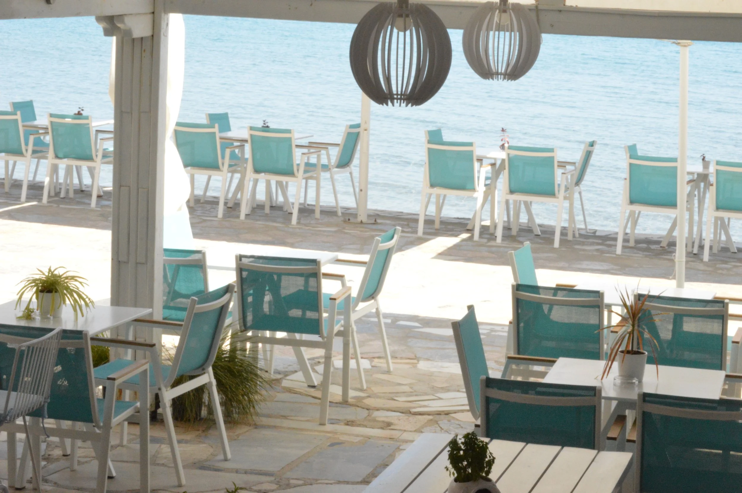 Outdoor seating area at a restaurant or cafe by a body of water, with white tables and blue chairs, and two hanging spherical light fixtures.