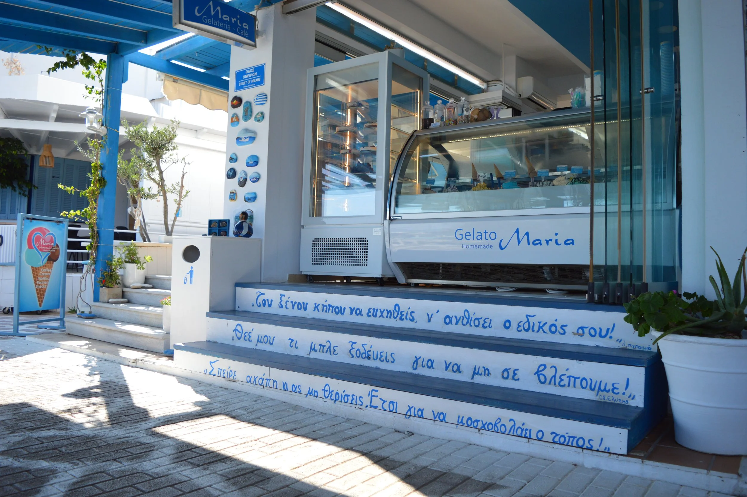 An outdoor Greek gelato shop called 'Gelato Maria' with a white and blue color scheme. The shop has a glass display case full of gelato, stairs with Greek writing on the risers, and a signboard with an ice cream cone logo outside.
