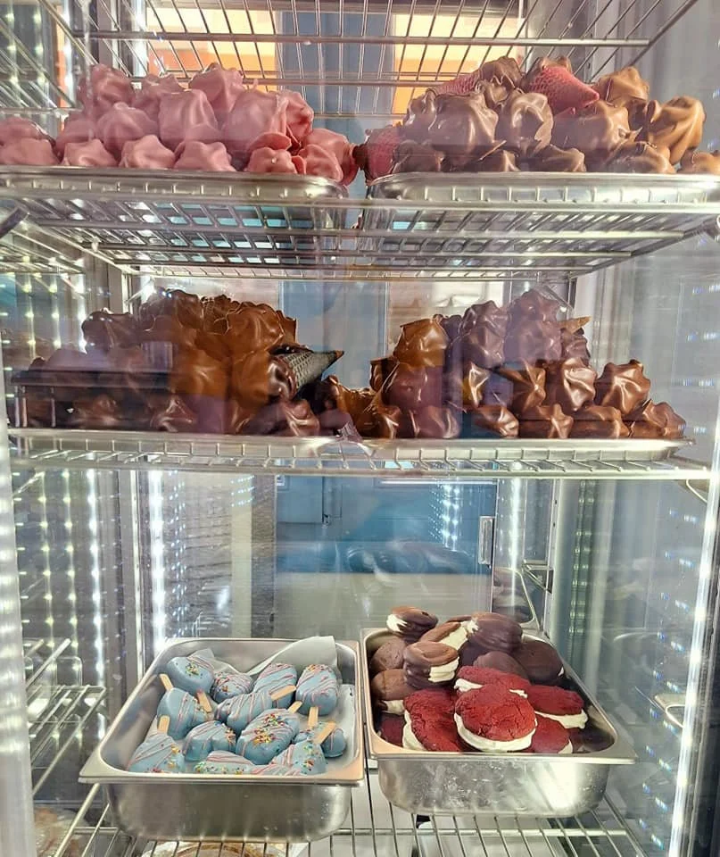 Cold storage refrigerator containing various ice cream treats on three shelves. The top shelf has pink and purple ice cream cones, the middle shelf has chocolate-covered ice cream bars and cones, and the bottom shelf has chocolate and red velvet ice cream sandwiches with popsicle sticks and ice cream cookies with ice cream filling.