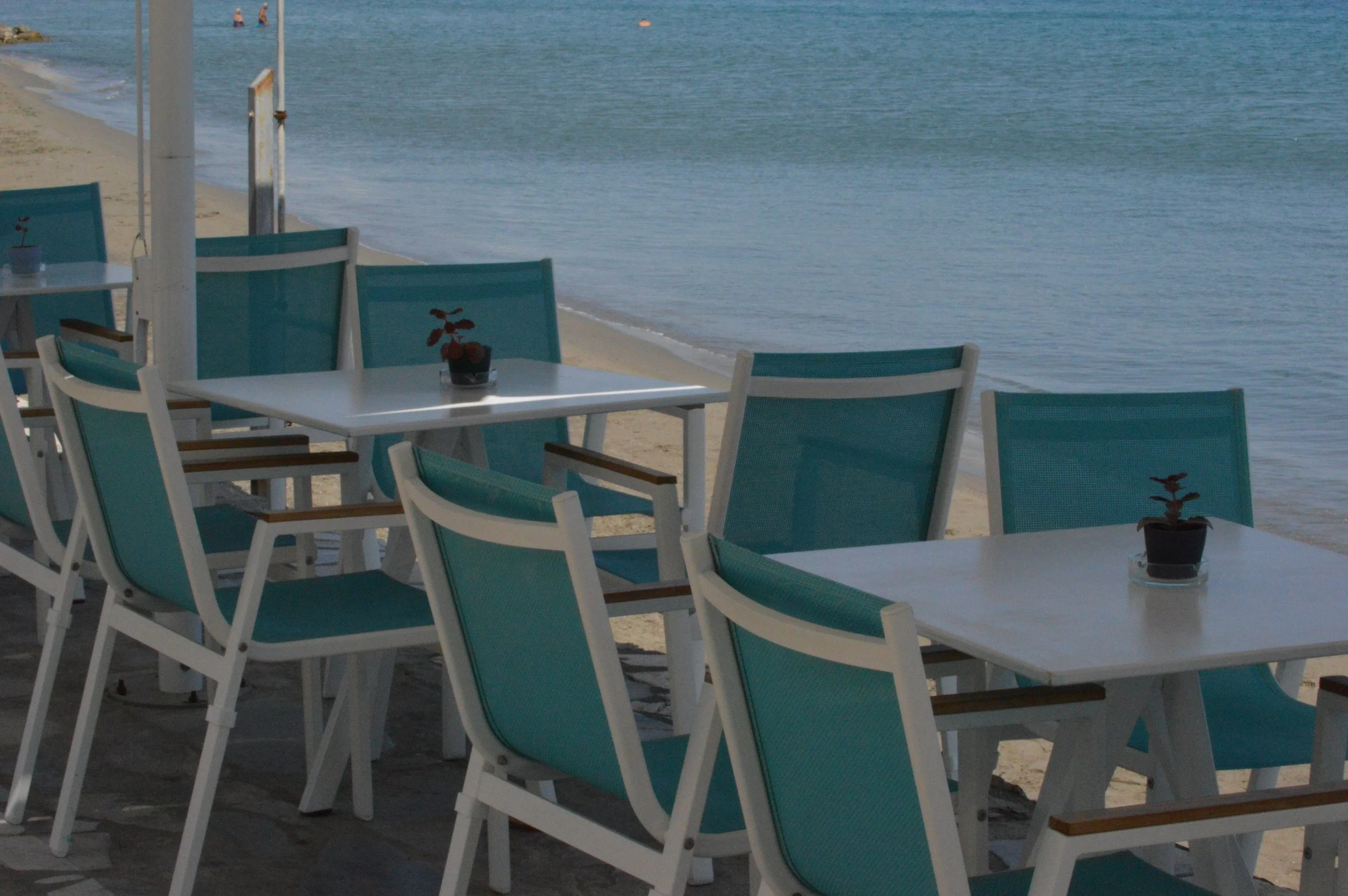 Gelateria Maria - Empty outdoor seating area with white tables and teal chairs on a sandy beach, overlooking calm ocean water.