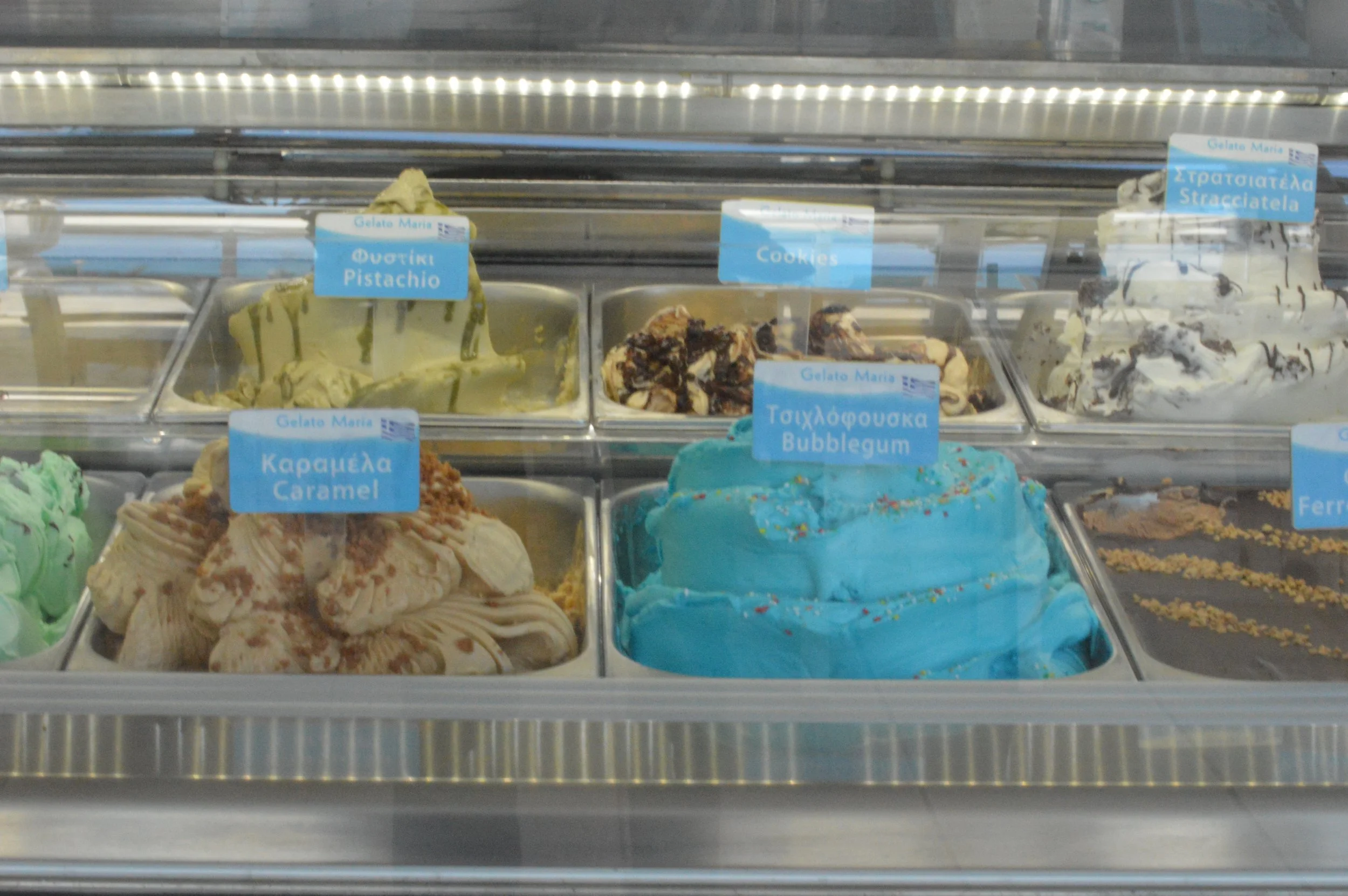 Gelateria Maria - Selection of gelato flavors in a gelato shop, including pistachio, caramel, cookies, bubblegum, and stracciatella, with labeling in Greek and English.
