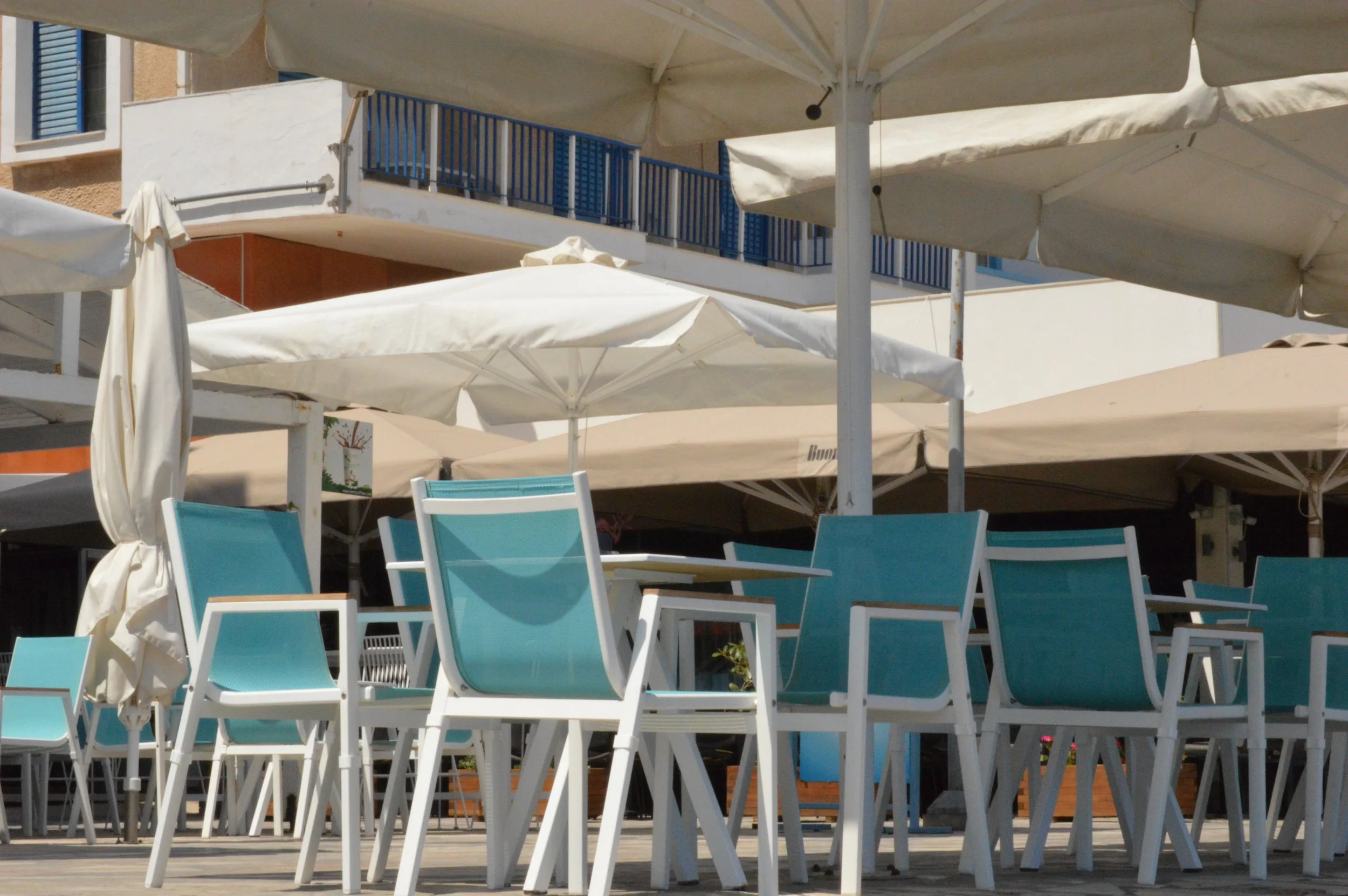Empty outdoor seating area with turquoise chairs and closed umbrellas at a beachside restaurant.