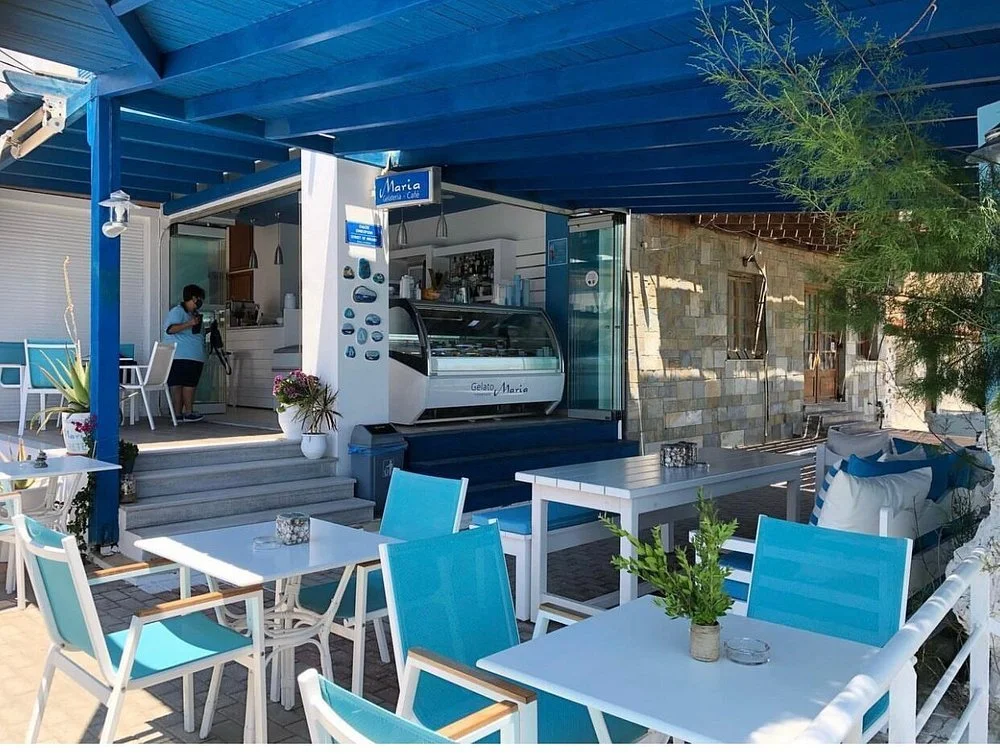 Gelateria Maria - Outdoor seating area of a cafe with white tables, blue chairs, and potted plants, featuring a gelato display case and a woman taking a photo inside.