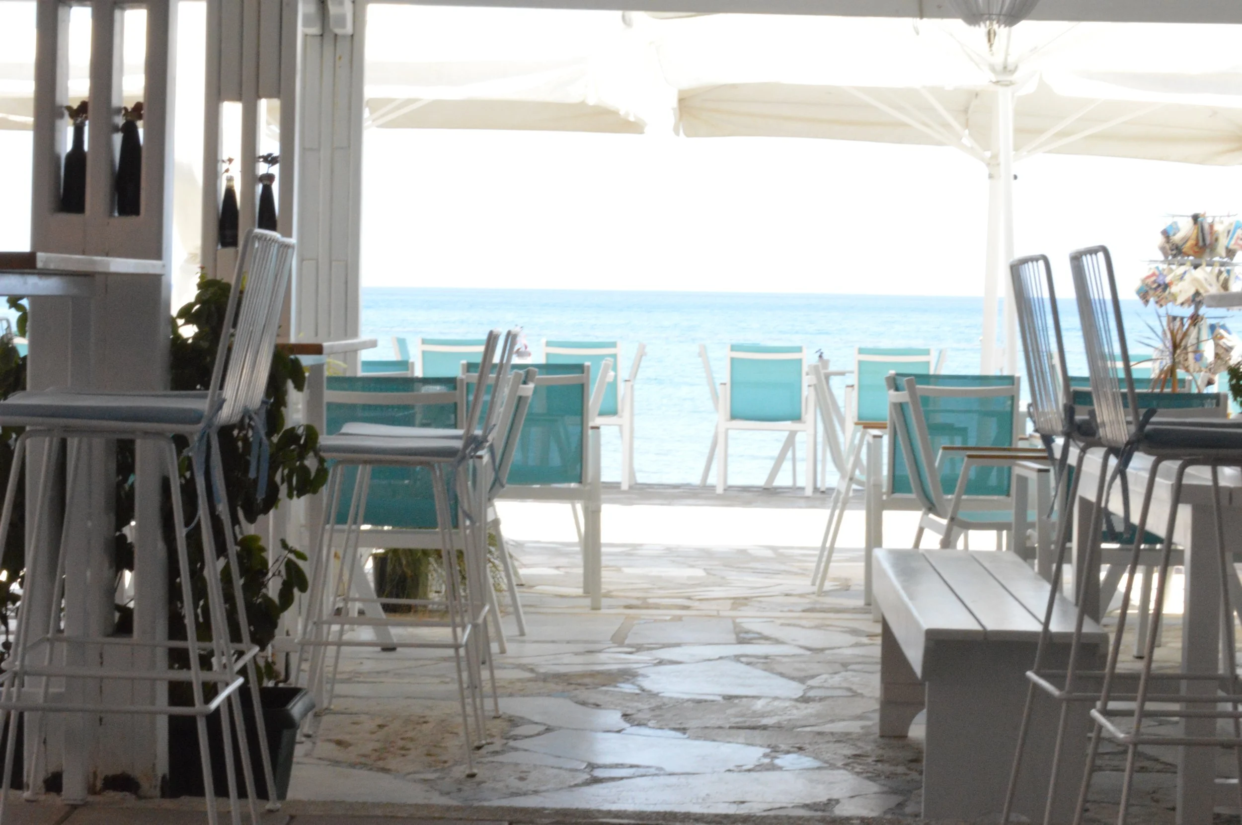 Gelateria Maria - View of a seaside restaurant or cafe with turquoise chairs and tables, white wooden furniture, and a white canopy providing shade, overlooking the ocean.